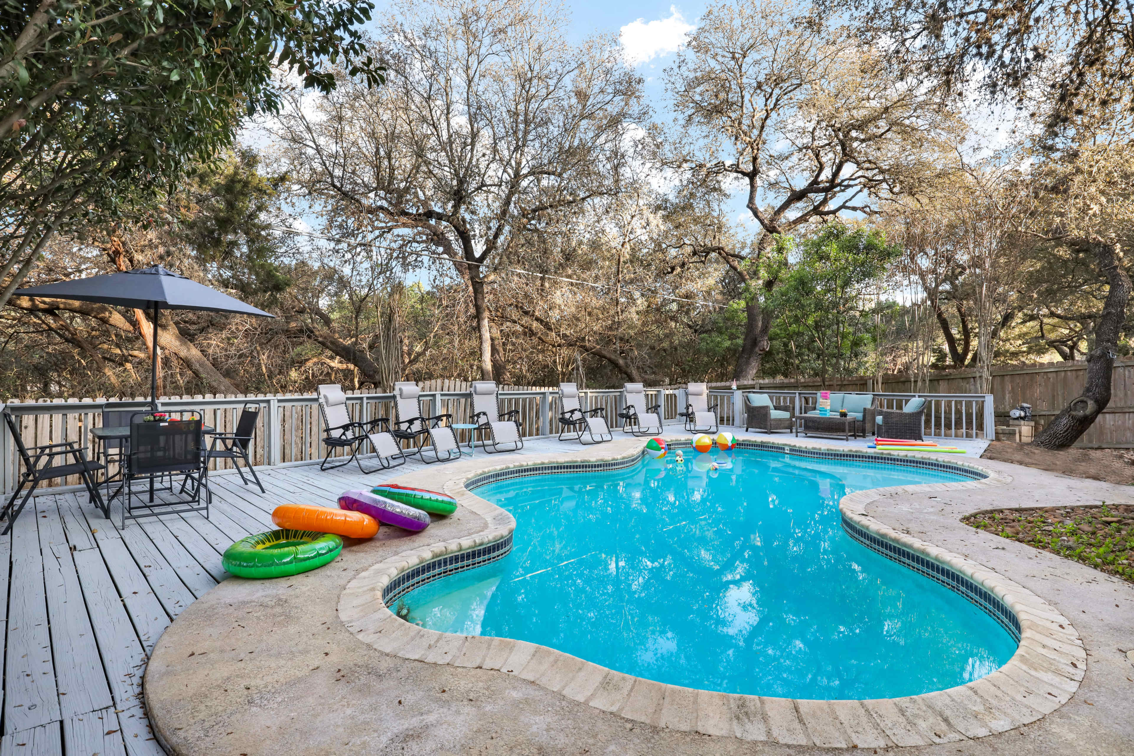 A swimming pool with colorful floats is surrounded by a wooden deck and lawn chairs under an umbrella, situated in a wooded outdoor area.