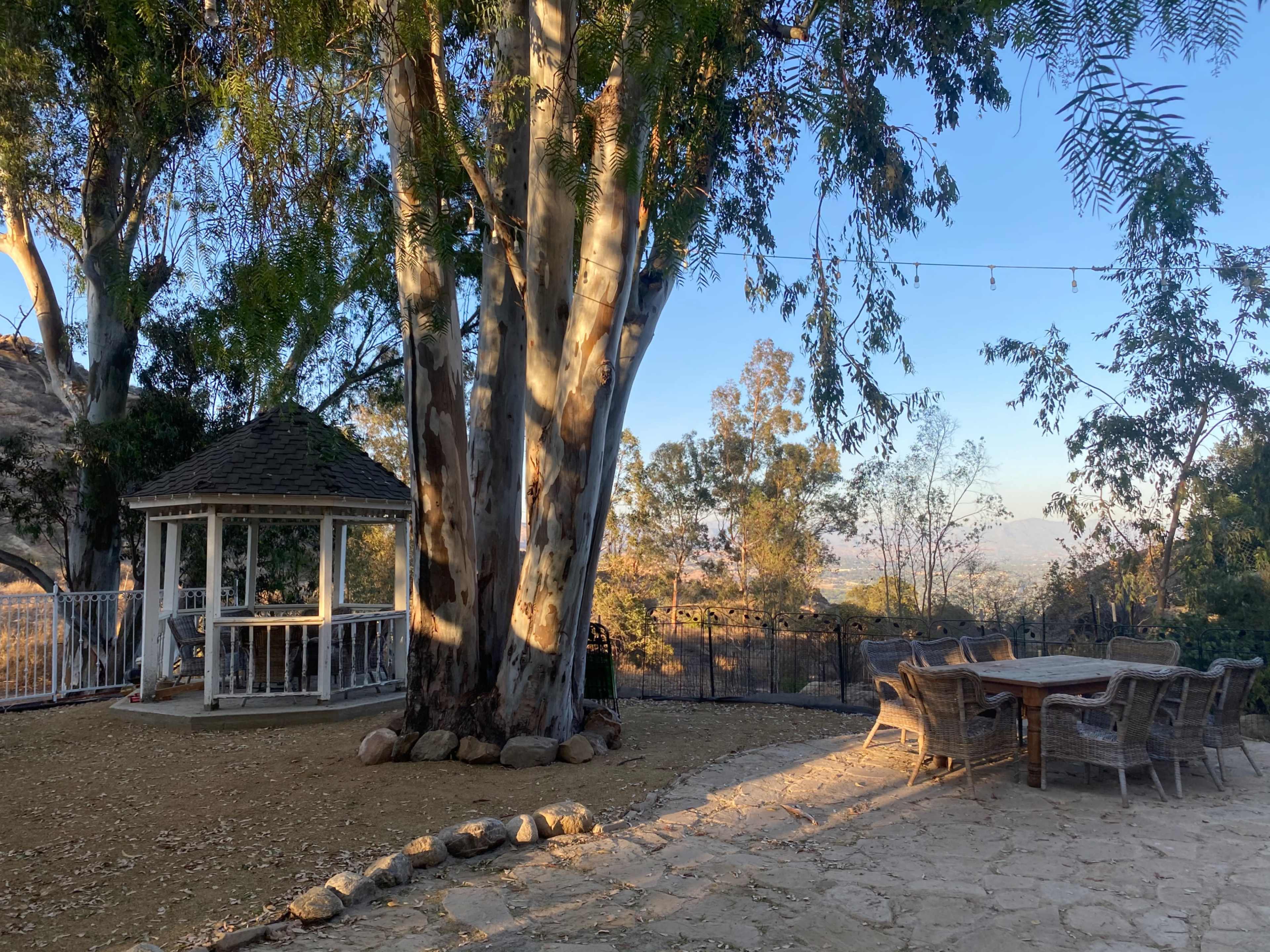 A gazebo stands beside a stone path leading to a dining table, surrounded by trees with a distant view of the landscape.