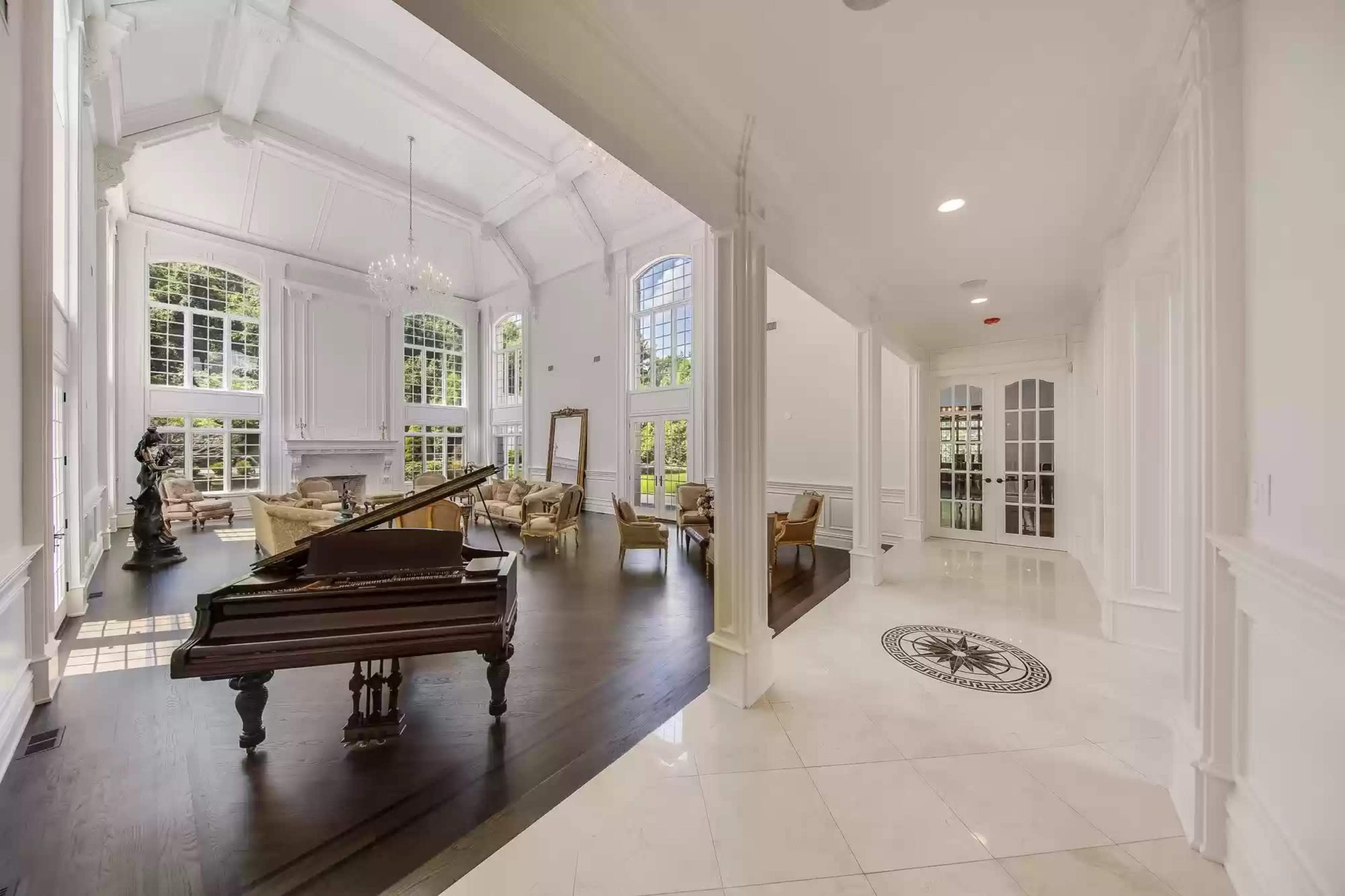 A large, elegant foyer opens into a spacious living area featuring a grand piano, high ceilings, and ample natural light streaming through large windows.
