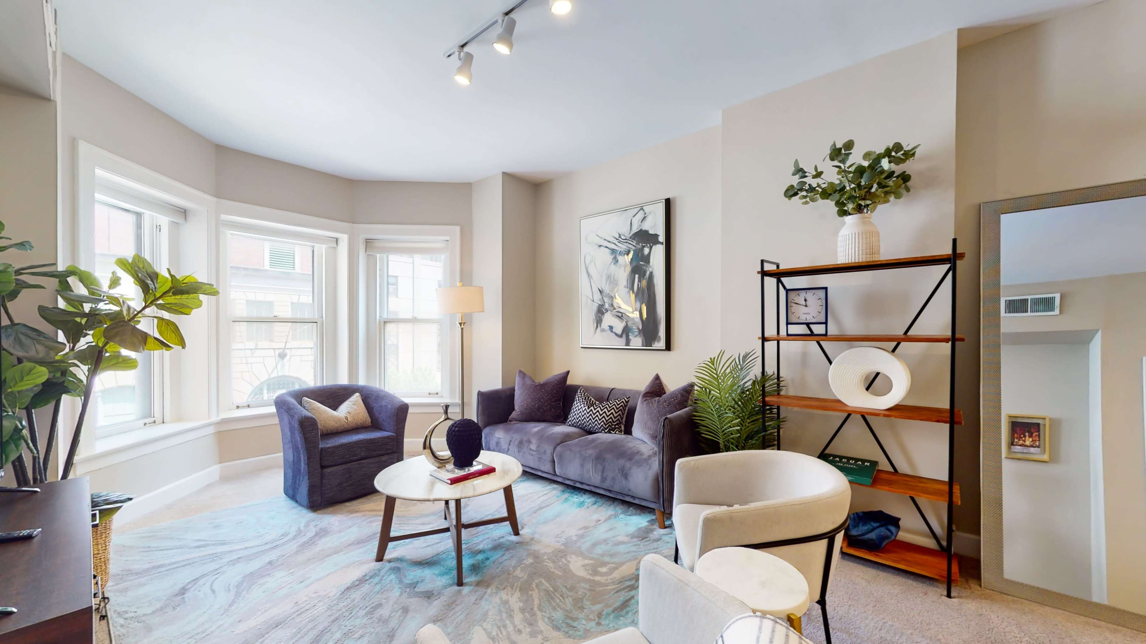 The image shows a modern living room featuring a gray sofa, a round coffee table, a decorative shelving unit, and large windows allowing natural light.