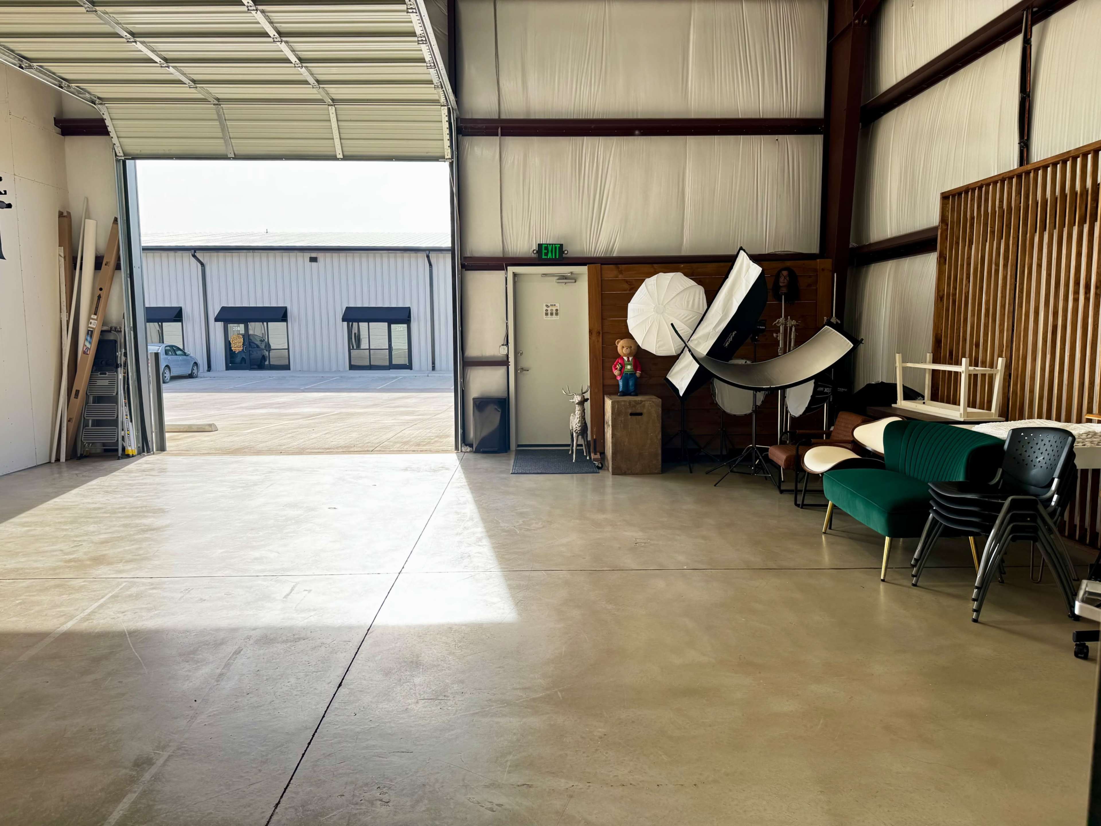 The image shows the interior of a large warehouse with an open door, featuring various pieces of furniture and photography equipment scattered throughout the space.