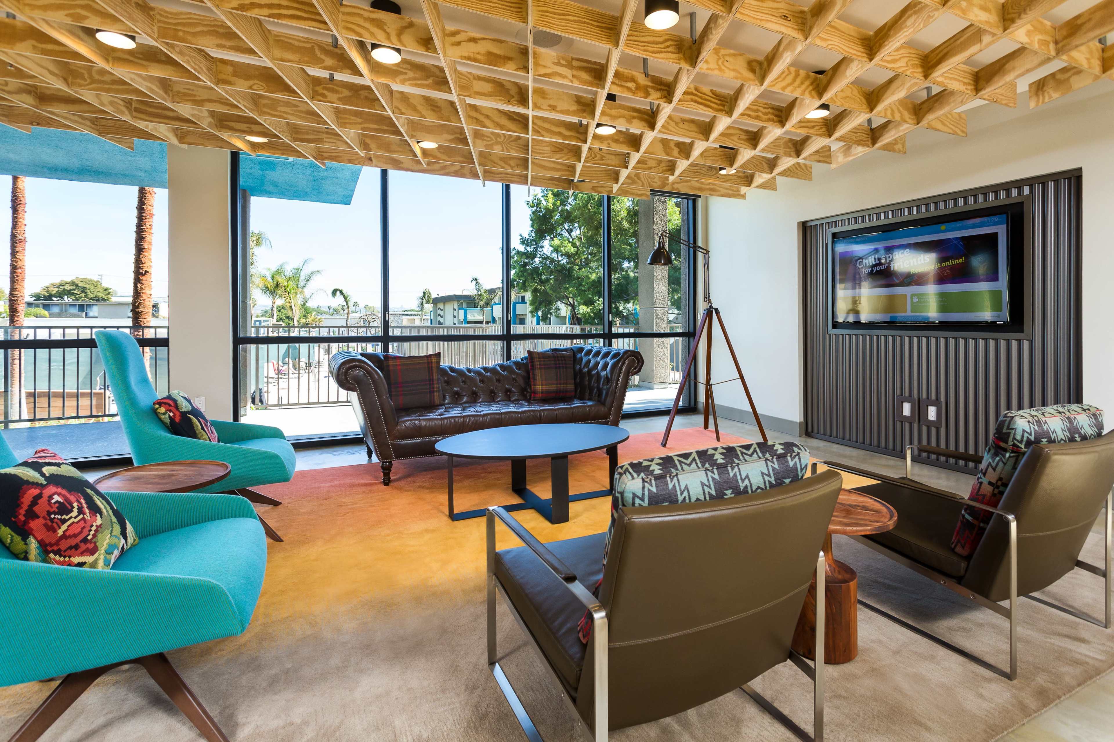 A modern lounge area featuring a leather sofa, colorful accent chairs, a round coffee table, and a large window with a view of palm trees.