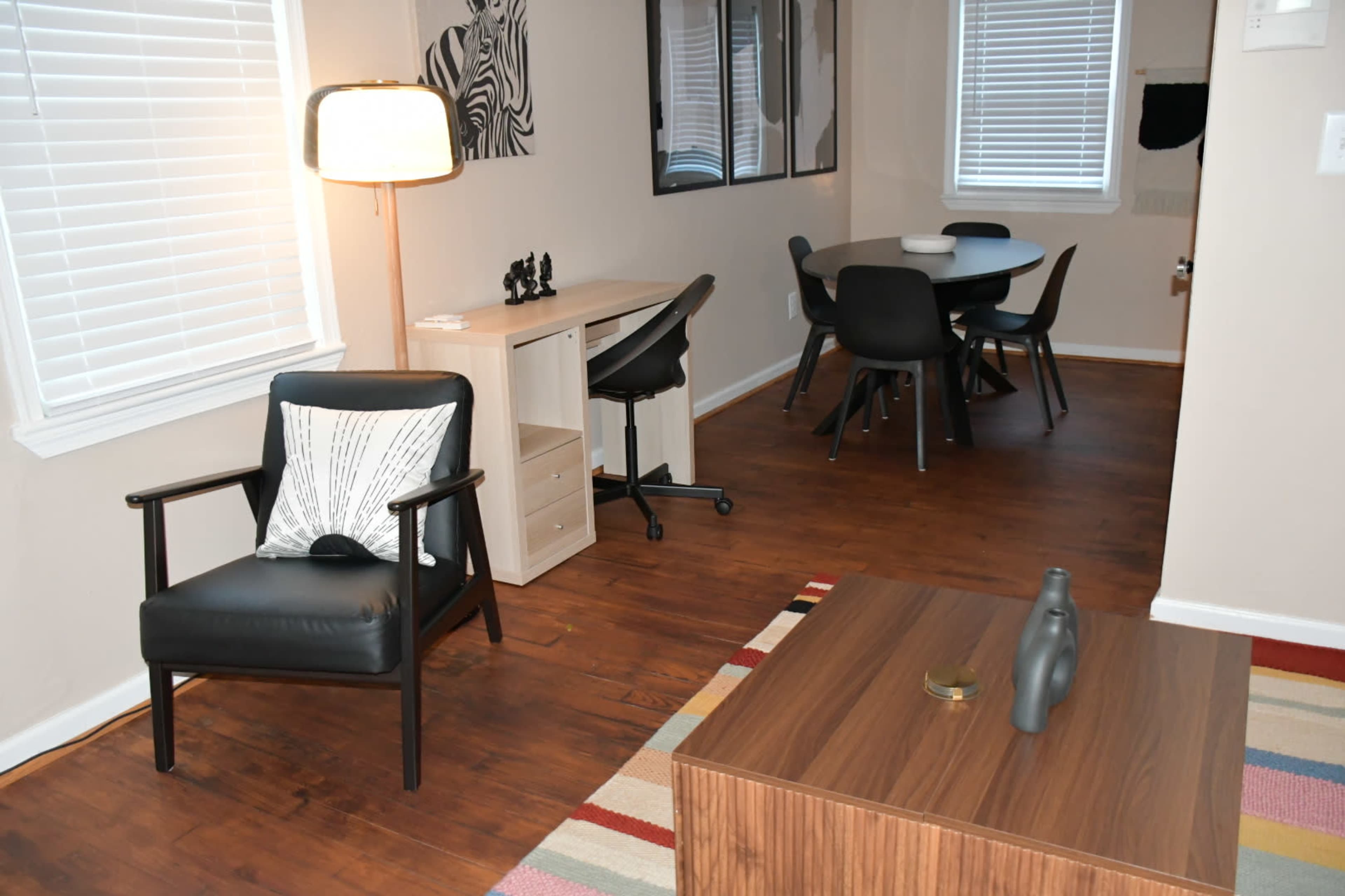 The image shows a well-lit living space featuring a black chair, a wooden table with chairs, and a coffee table on a patterned rug.