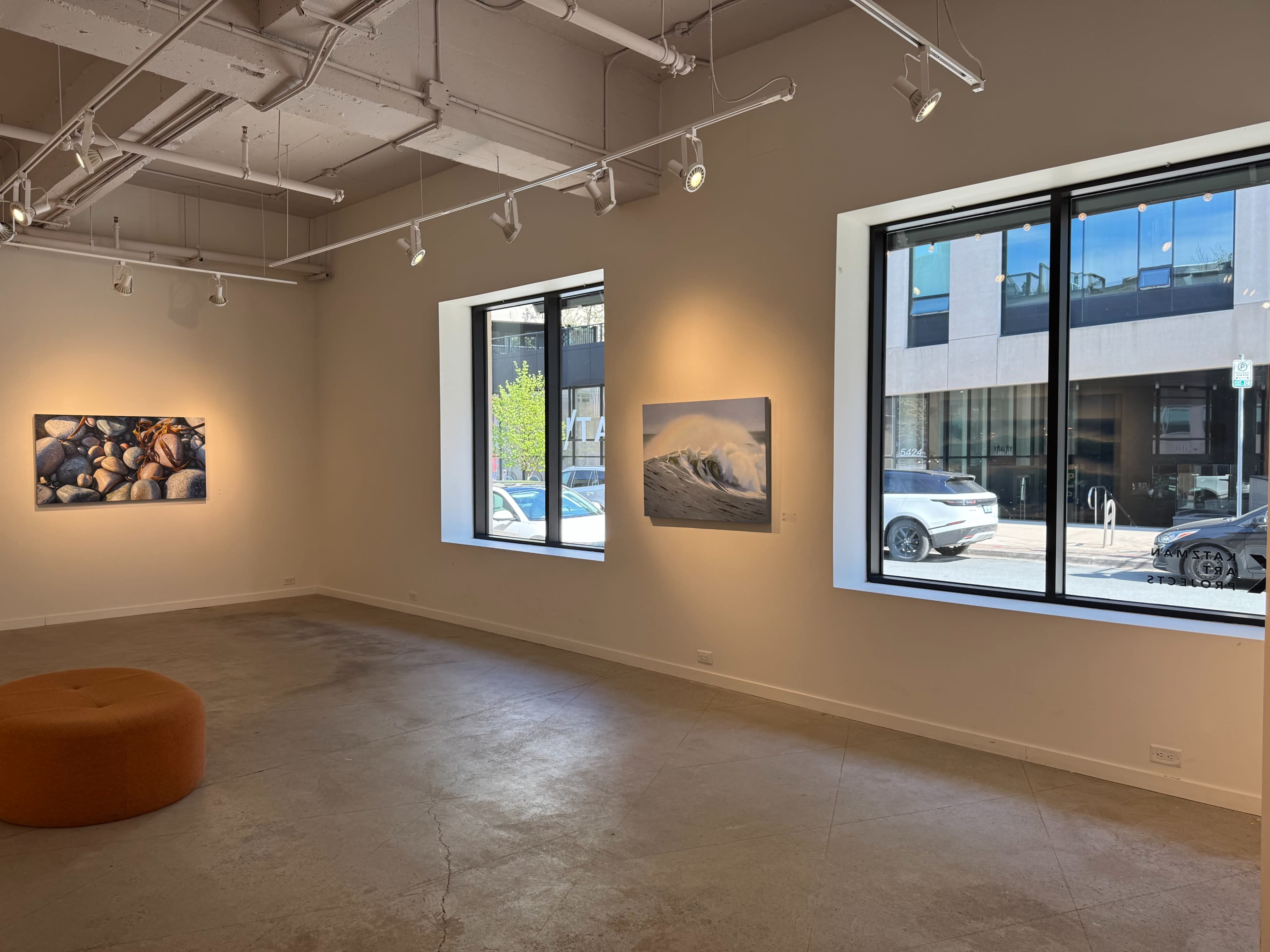 The image shows an art gallery with two large windows, one displaying a street view and the other featuring a photograph of rocks, alongside a wave image on the wall.