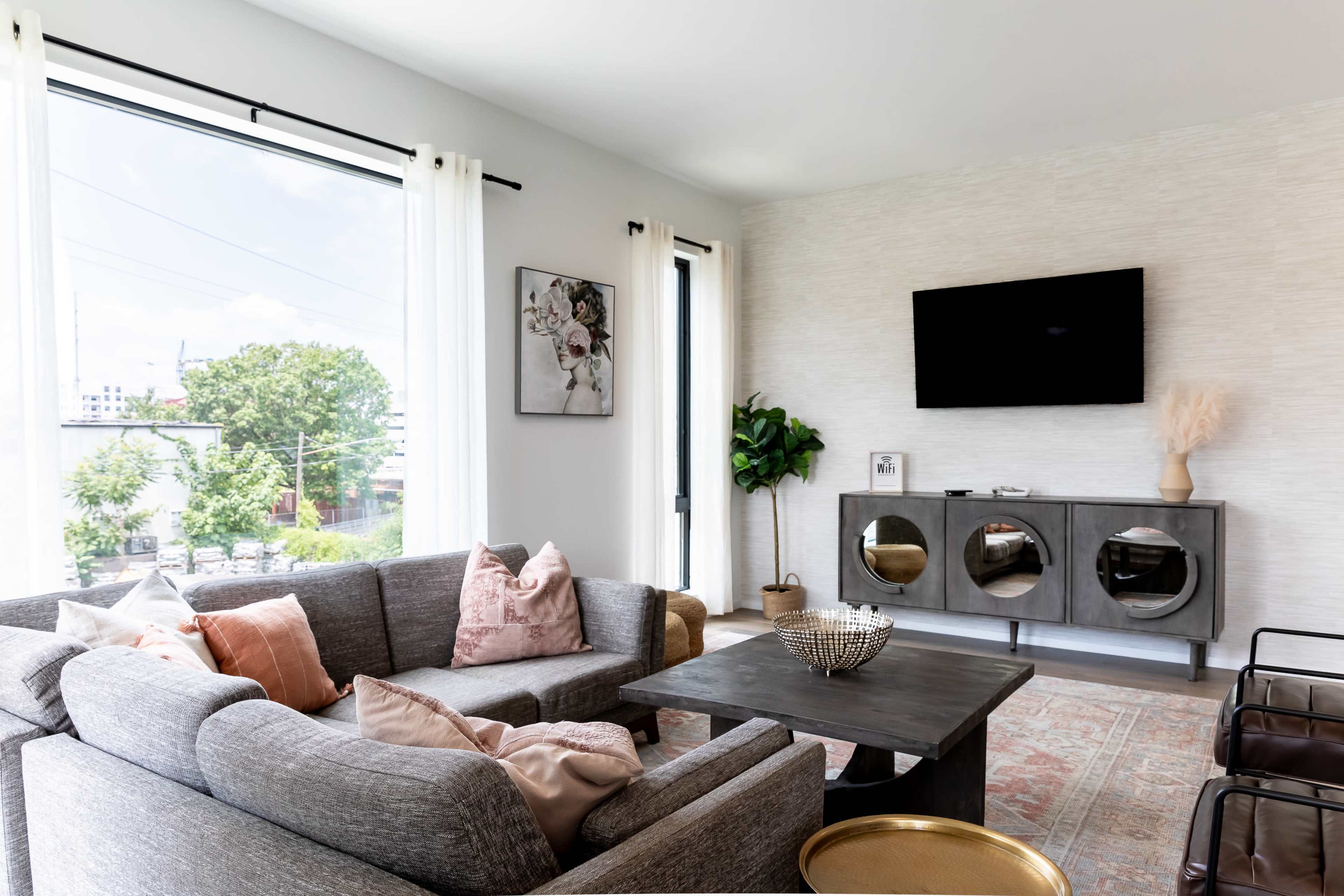 A modern living room features a gray sectional sofa, a wooden coffee table, and a wall-mounted television, with large windows allowing natural light to fill the space.