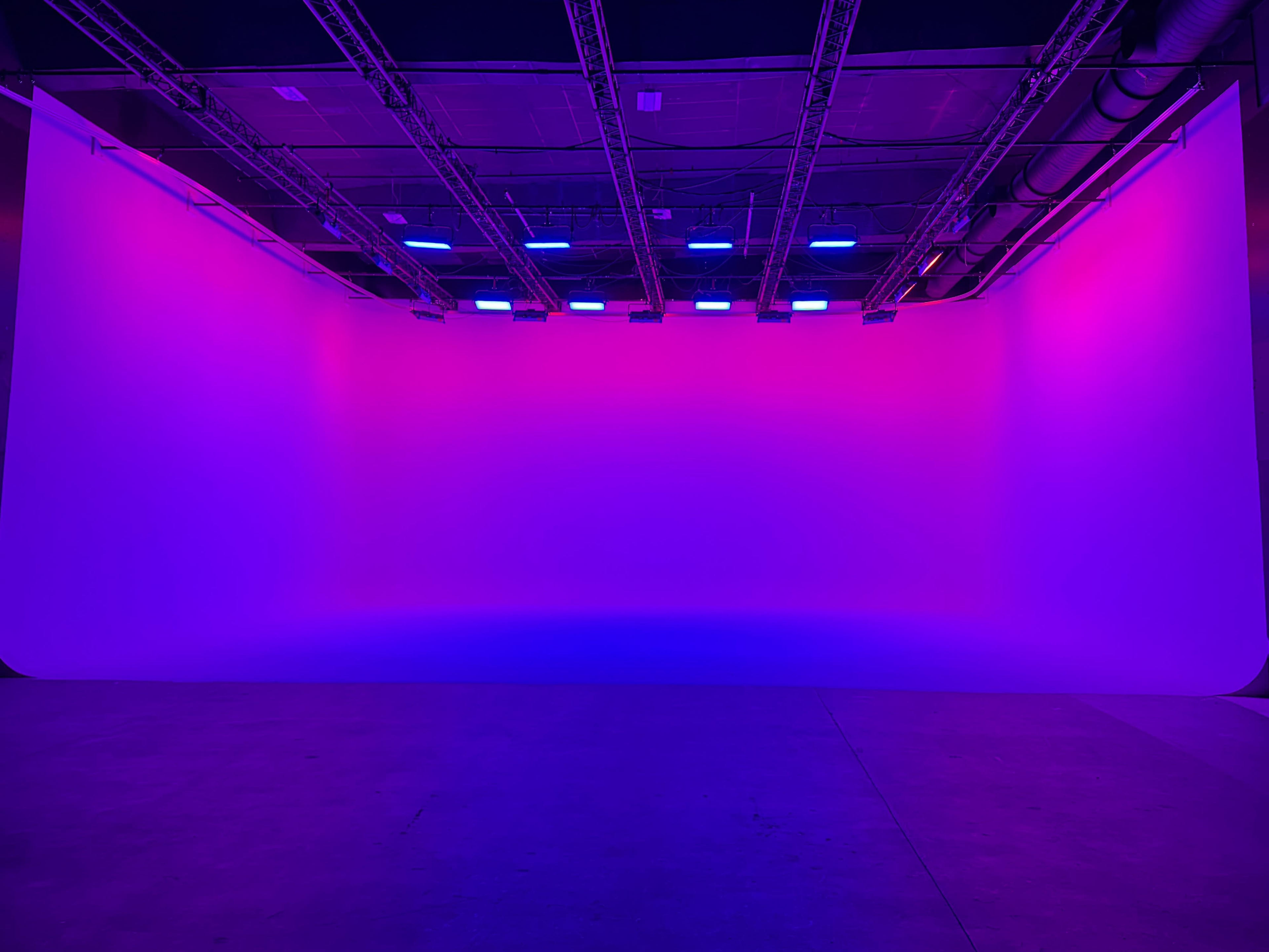 The image shows a large studio space with a backdrop illuminated in shades of pink and purple, featuring overhead lighting and visible structural elements.