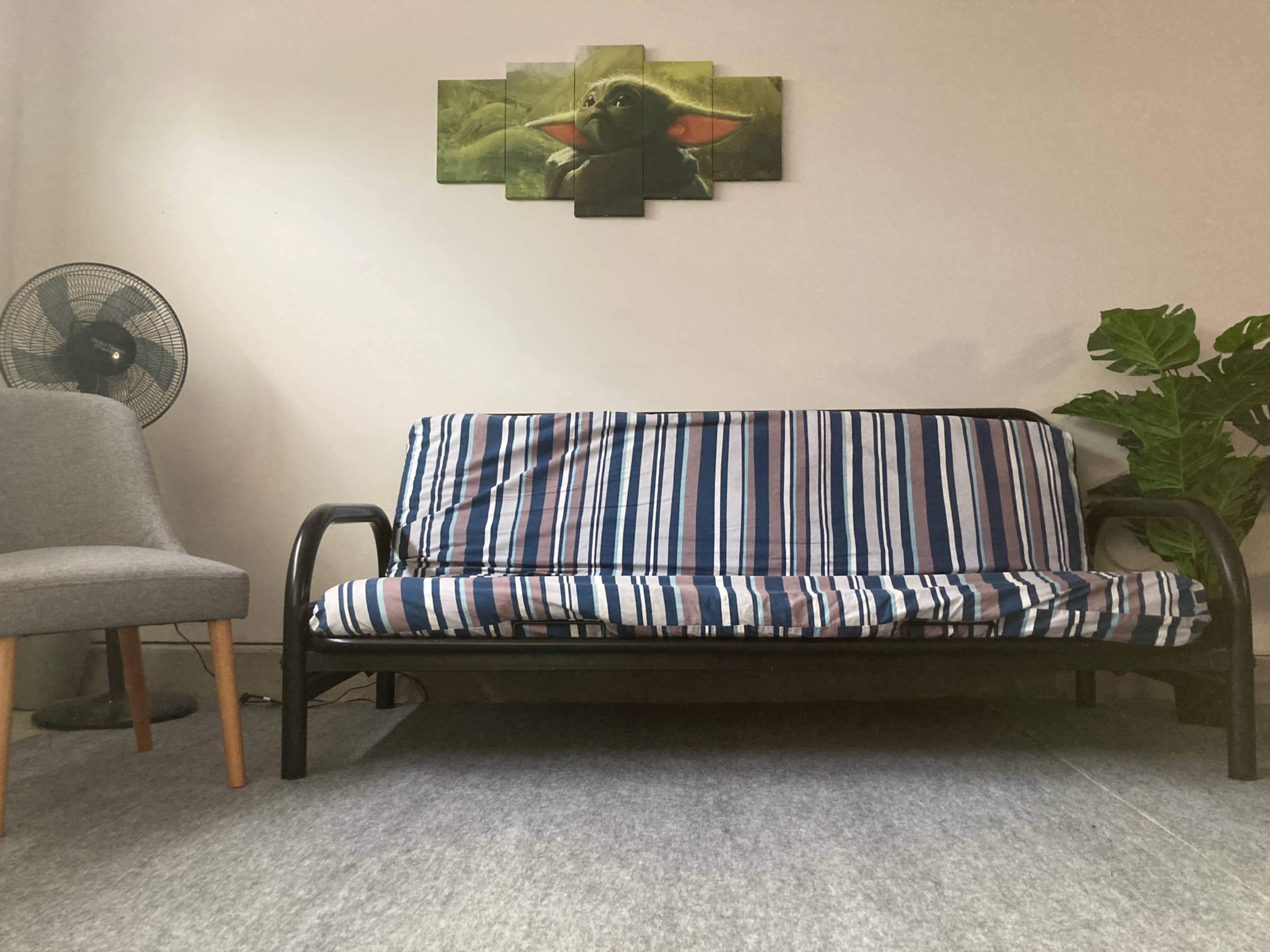The scene features a striped futon in a room with a green wall art piece of a character above it, a gray chair to the side, a fan, and a potted plant.