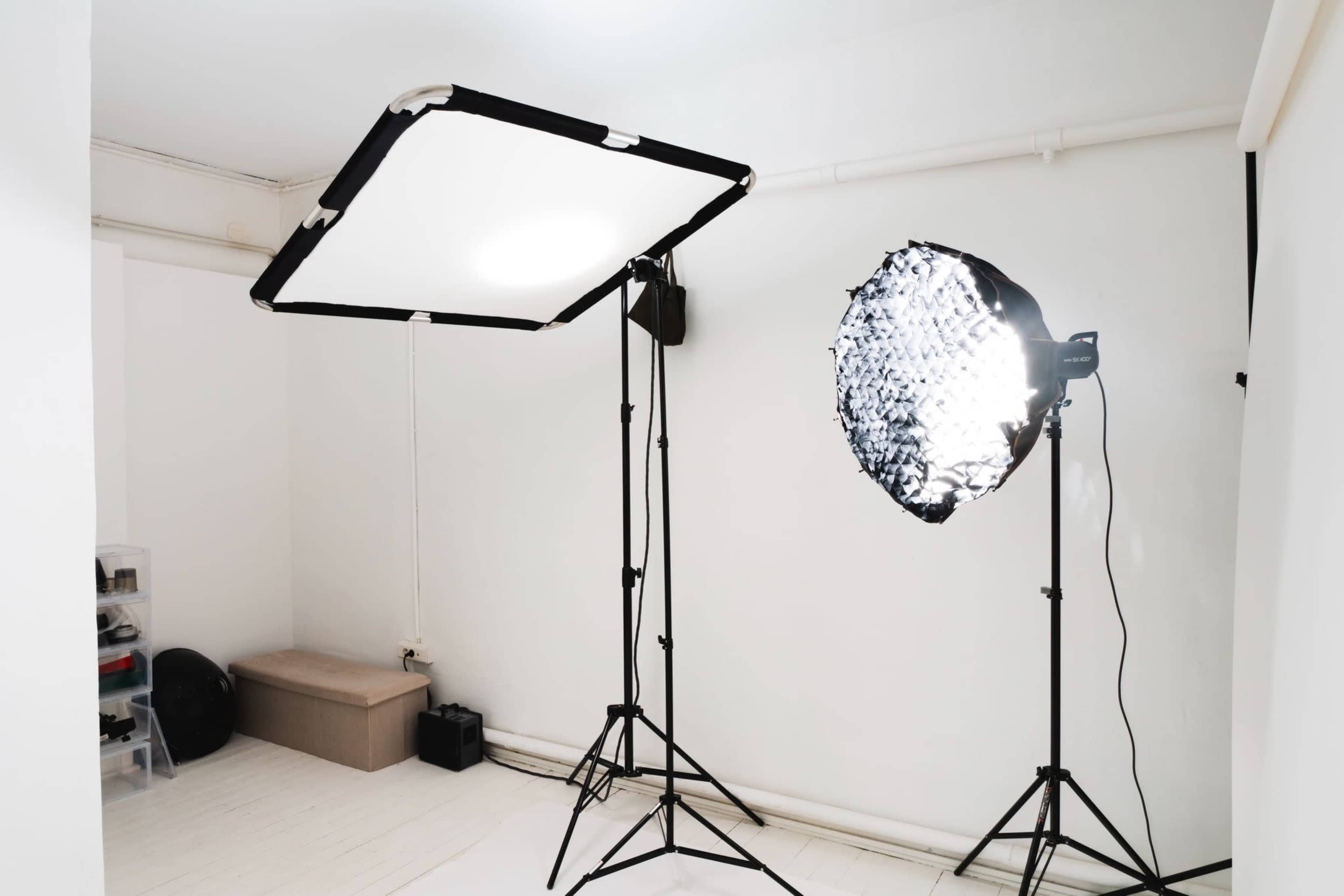 The image shows a photography studio setup featuring a large softbox lighting and a round reflective light, both mounted on stands, against a white wall.