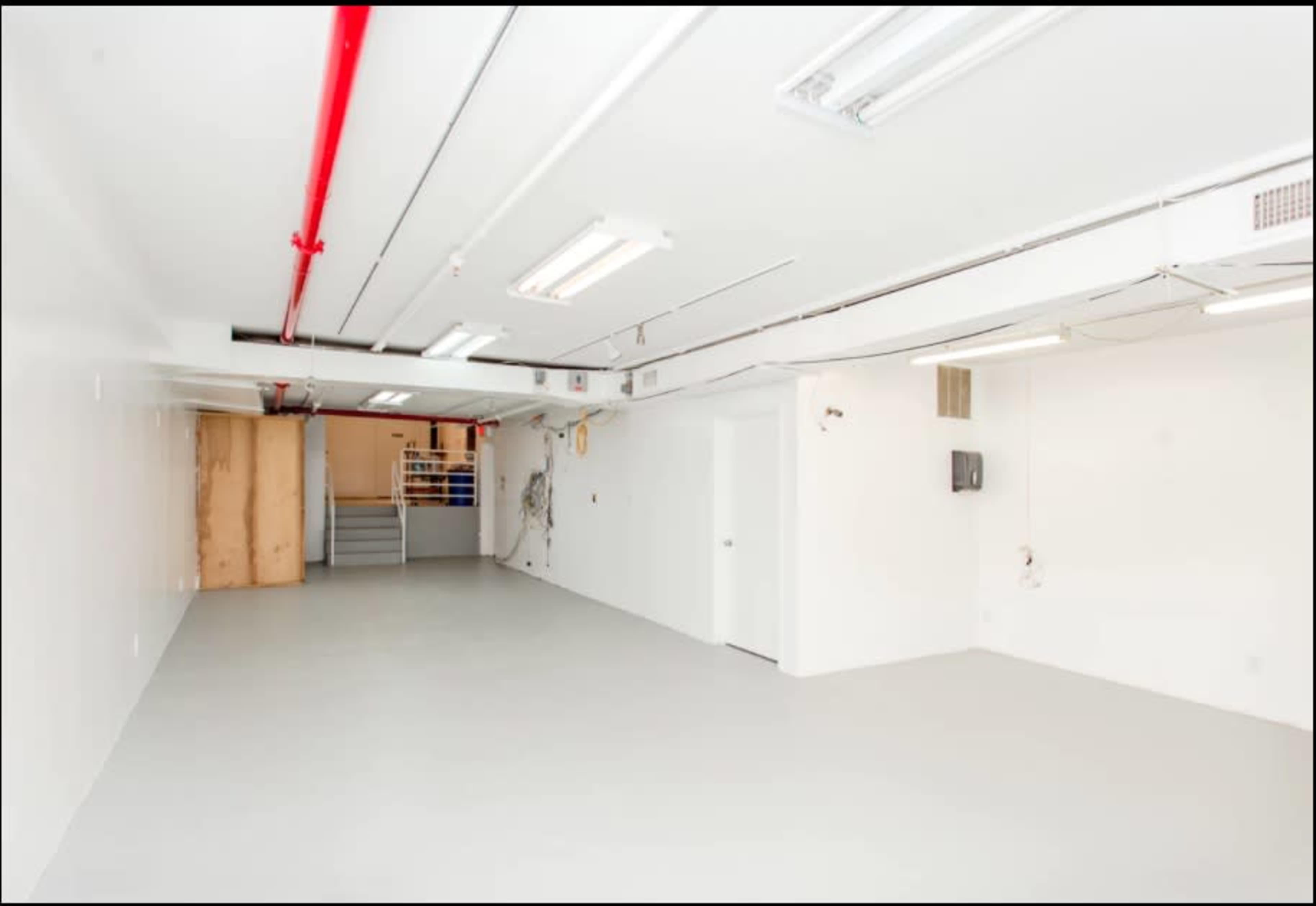 A large, empty basement space with white walls, fluorescent lighting, and a staircase in the background.