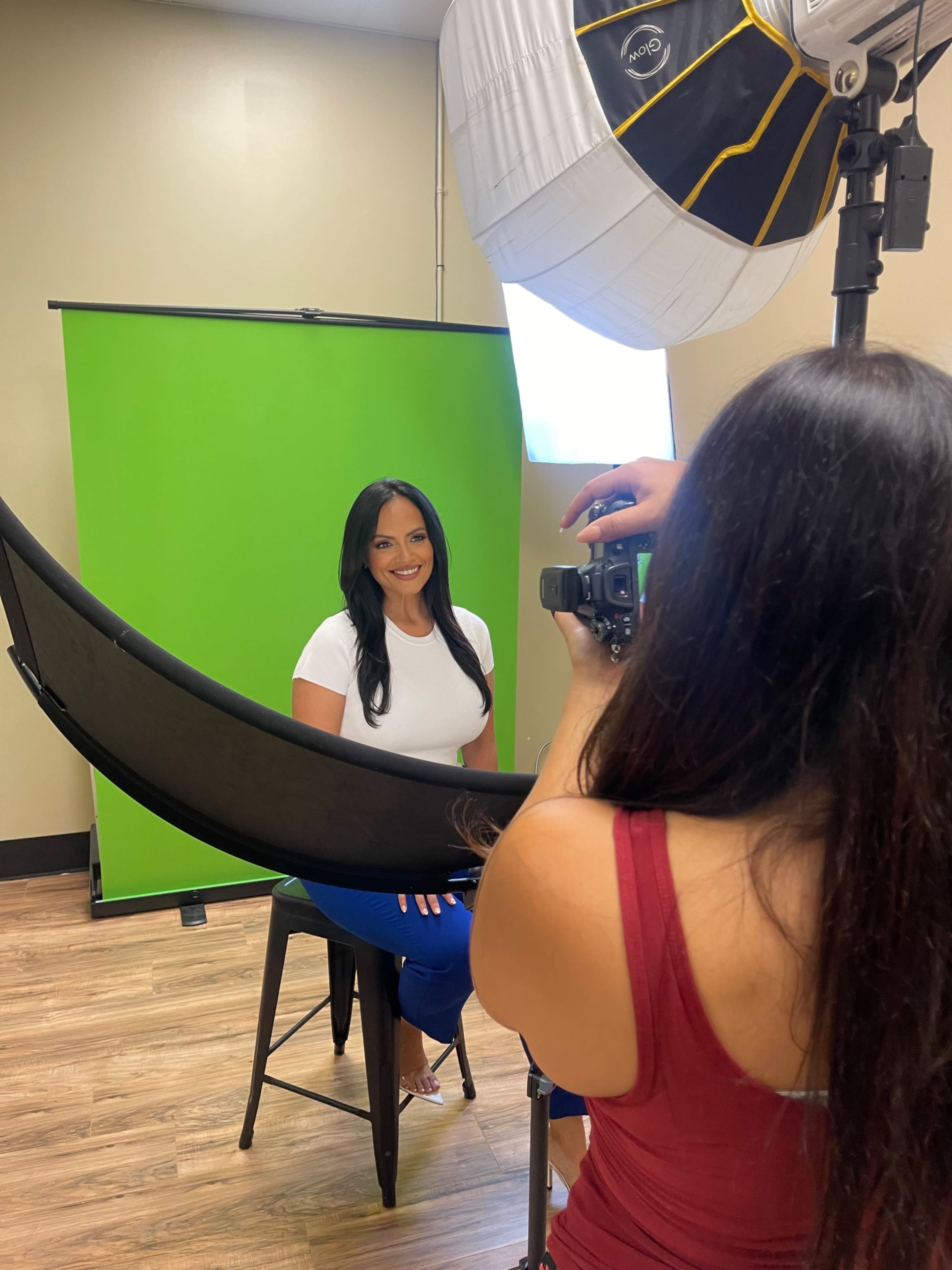A woman is seated on a stool in front of a green screen while another person takes her photo using a camera.