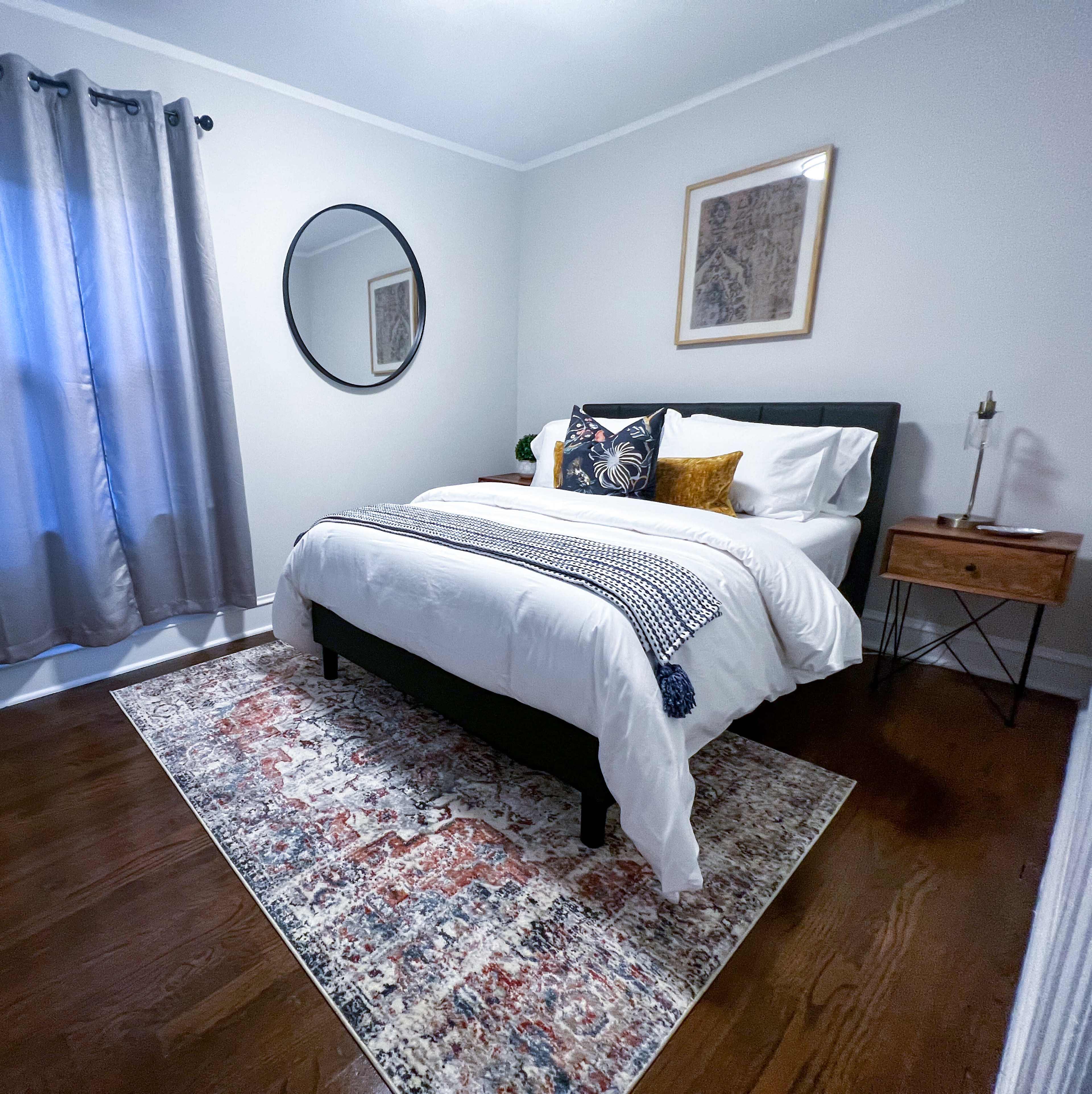 A neatly arranged bedroom featuring a bed with decorative pillows, a round mirror, a bedside table with a lamp, and a patterned rug on wooden flooring.
