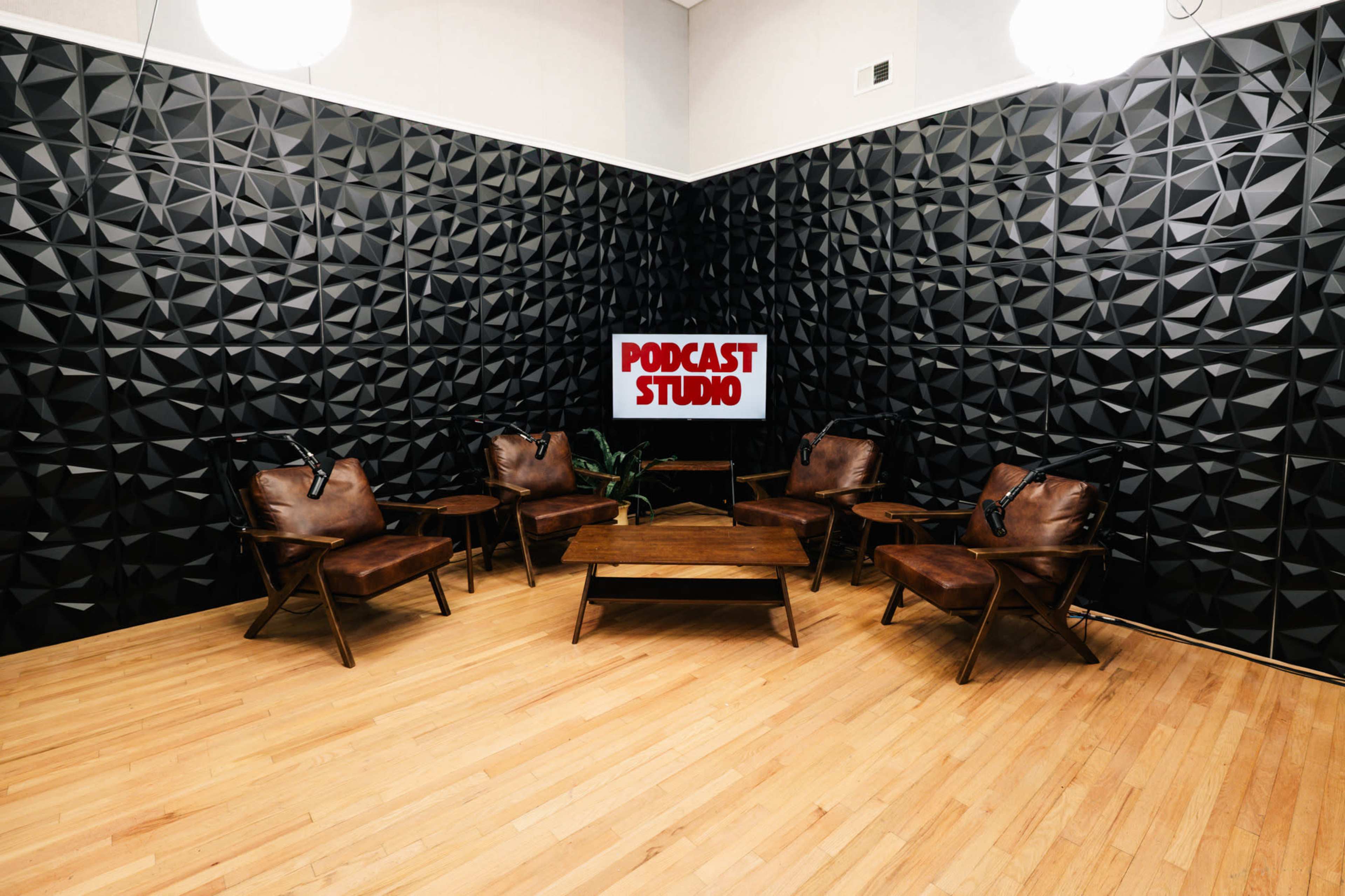 The image shows a podcast studio with textured black walls, wooden flooring, a table in the center, and leather chairs arranged around it, featuring a sign that reads "PODCAST STUDIO."