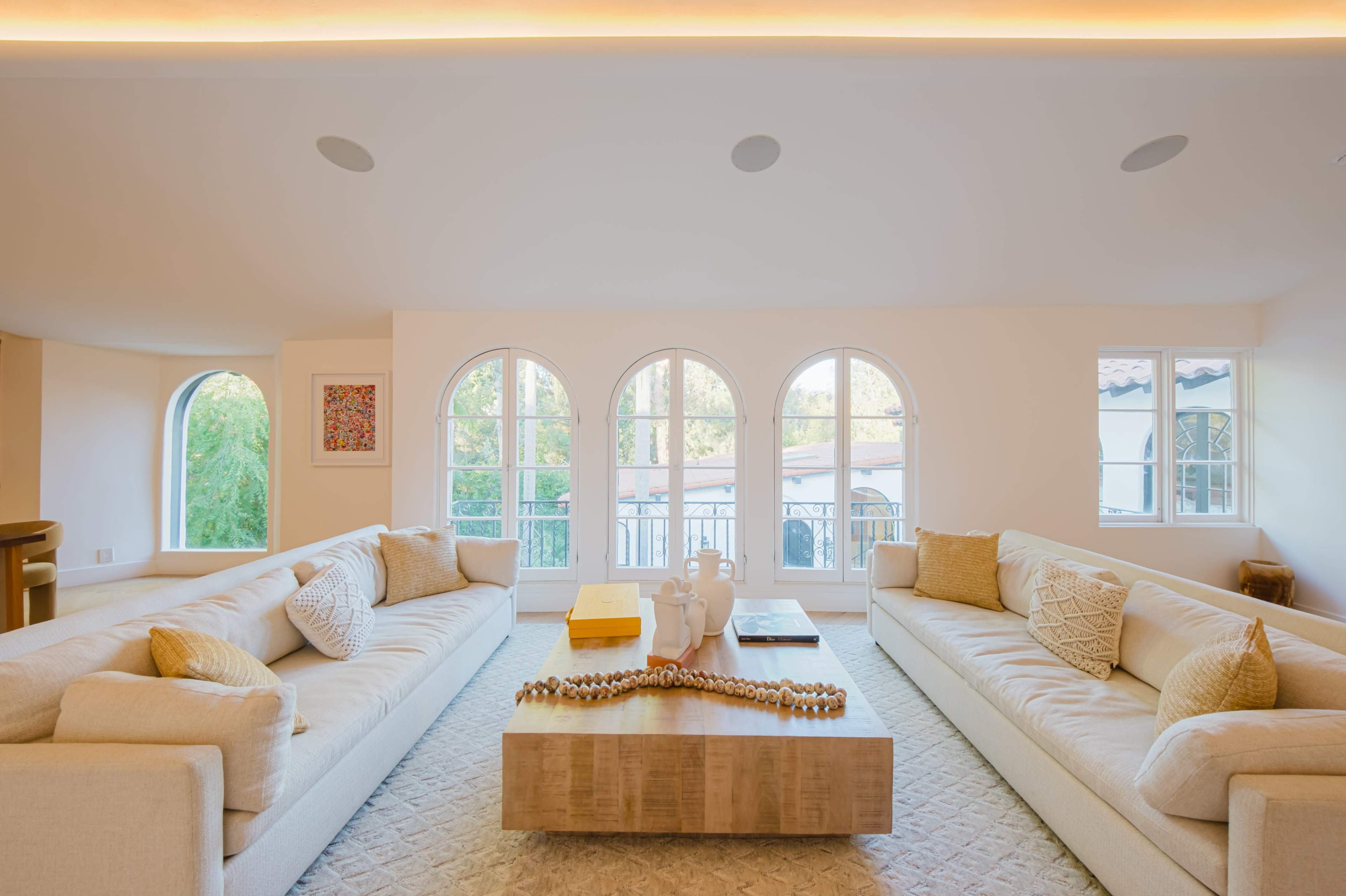 The image features a bright living room with two large sofas facing each other, a coffee table in the center, and arched windows that allow natural light to fill the space.