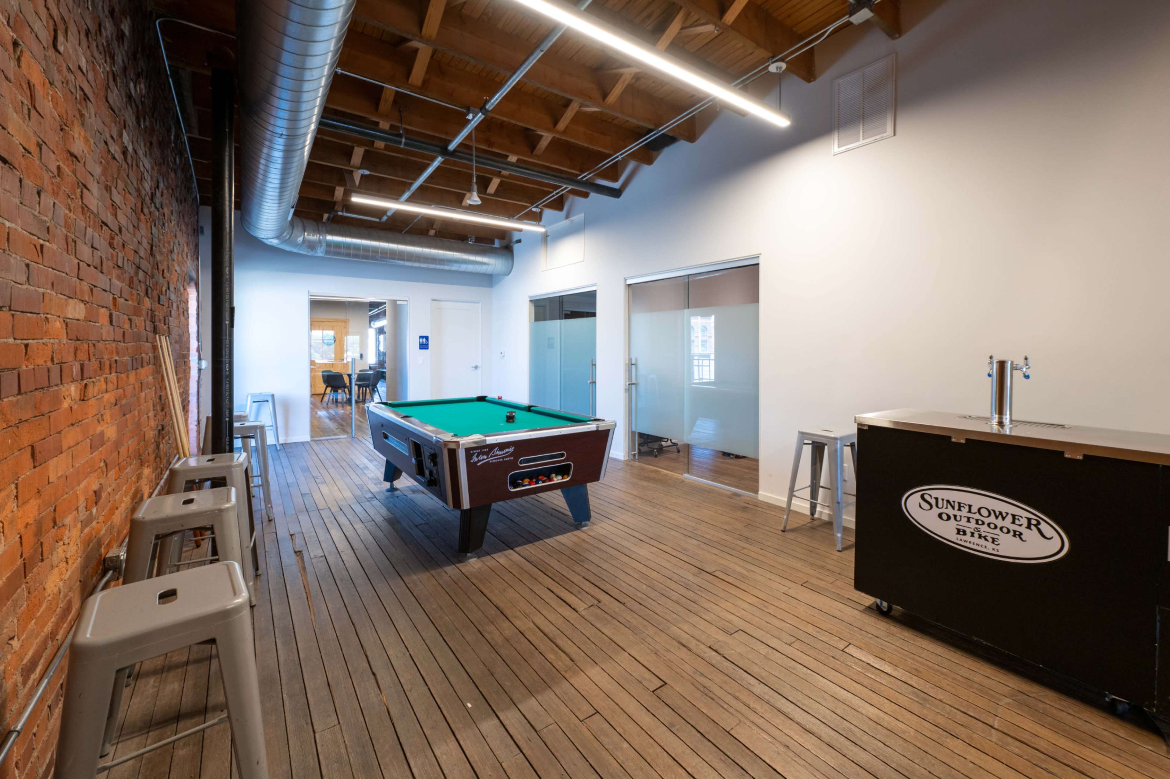 A pool table is centered in a modern workspace that features brick walls and wooden flooring, with a bar area nearby and glass-walled offices in the background.