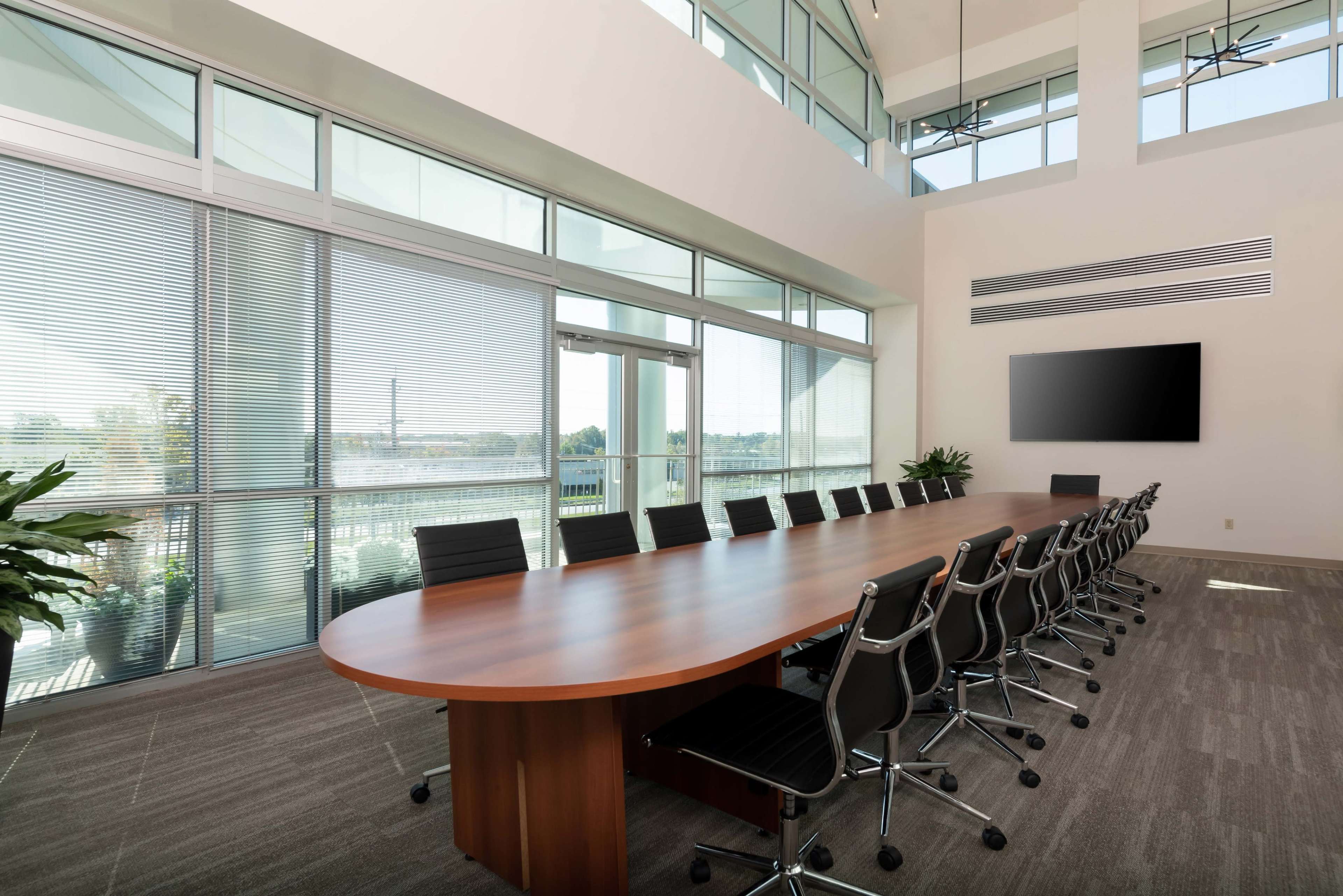 A spacious conference room featuring a long wooden table surrounded by black chairs, large windows with blinds, and a wall-mounted screen.