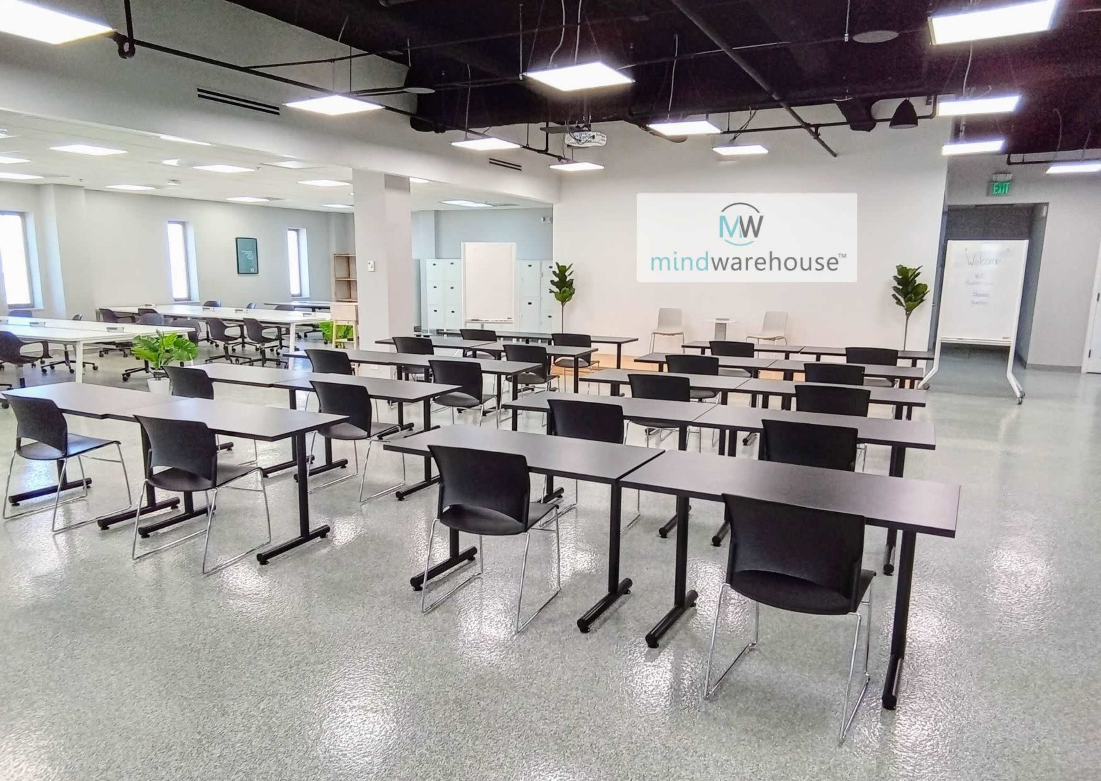 A spacious meeting room with rows of black chairs and tables, featuring a sign for "mindwarehouse" on the white wall.