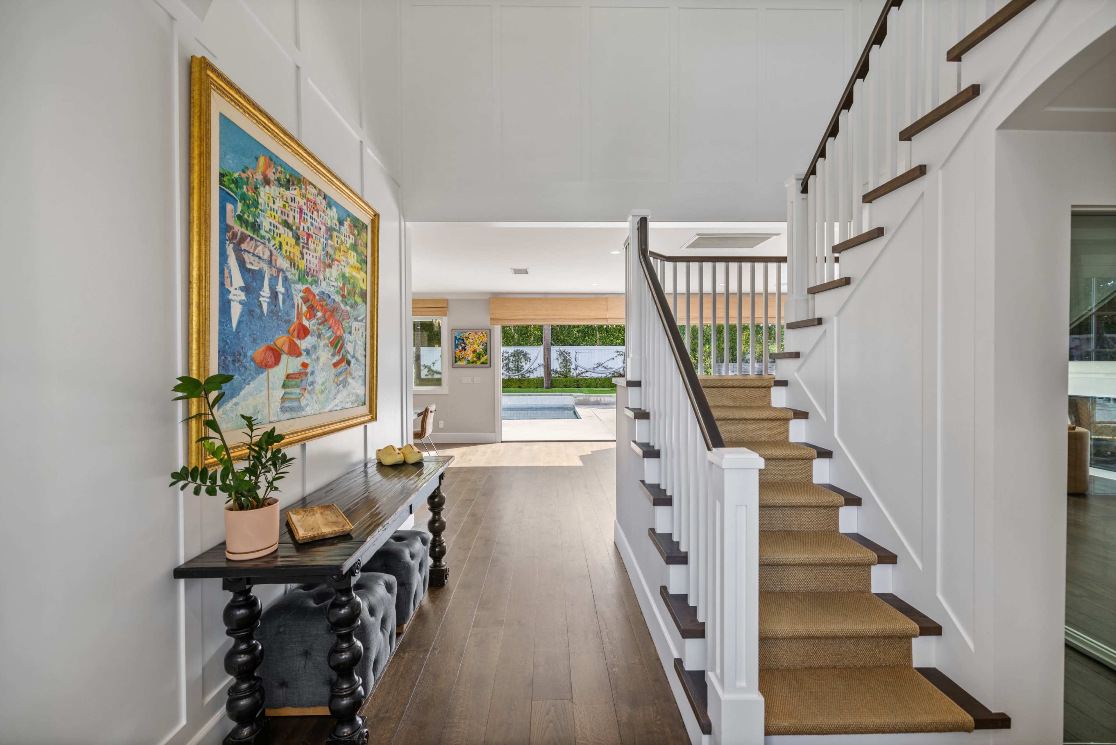 The image shows a well-lit hallway with a staircase, a colorful painting on the wall, and a dark console table with decorative items.