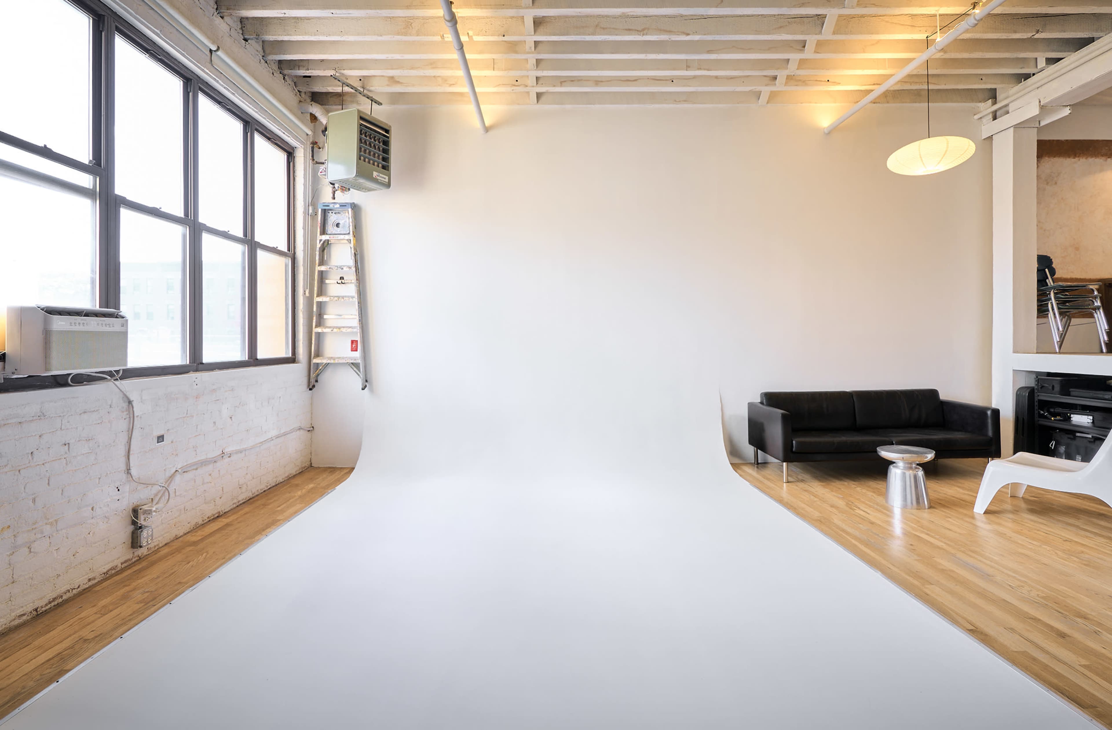 The image shows a minimalistic studio space with a white backdrop, wooden flooring, a black sofa, a small metallic table, and a ladder against the wall.
