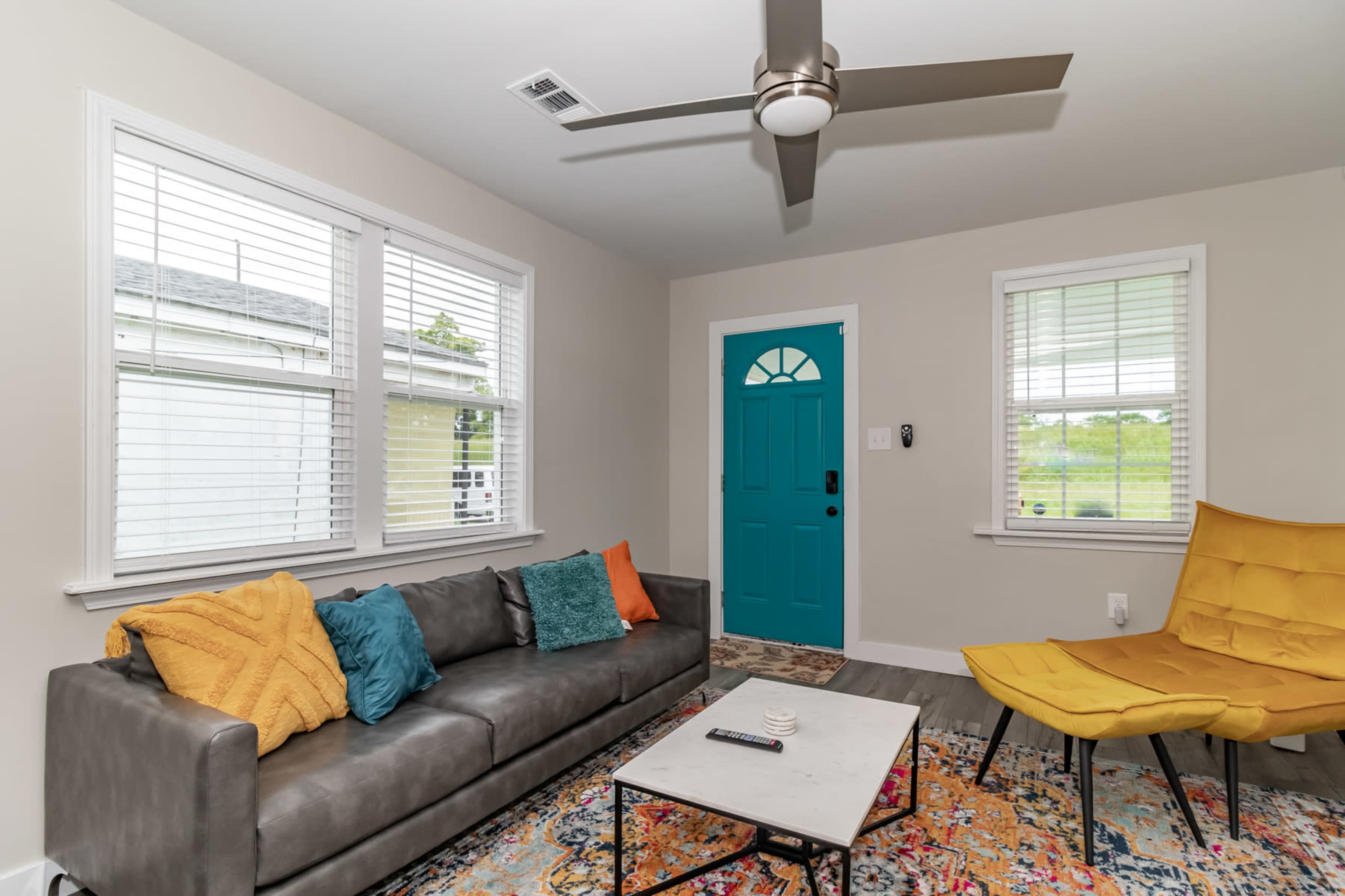 The living room features a gray sofa with colorful decorative pillows, a light coffee table, a vibrant area rug, and a door with a bright turquoise color.