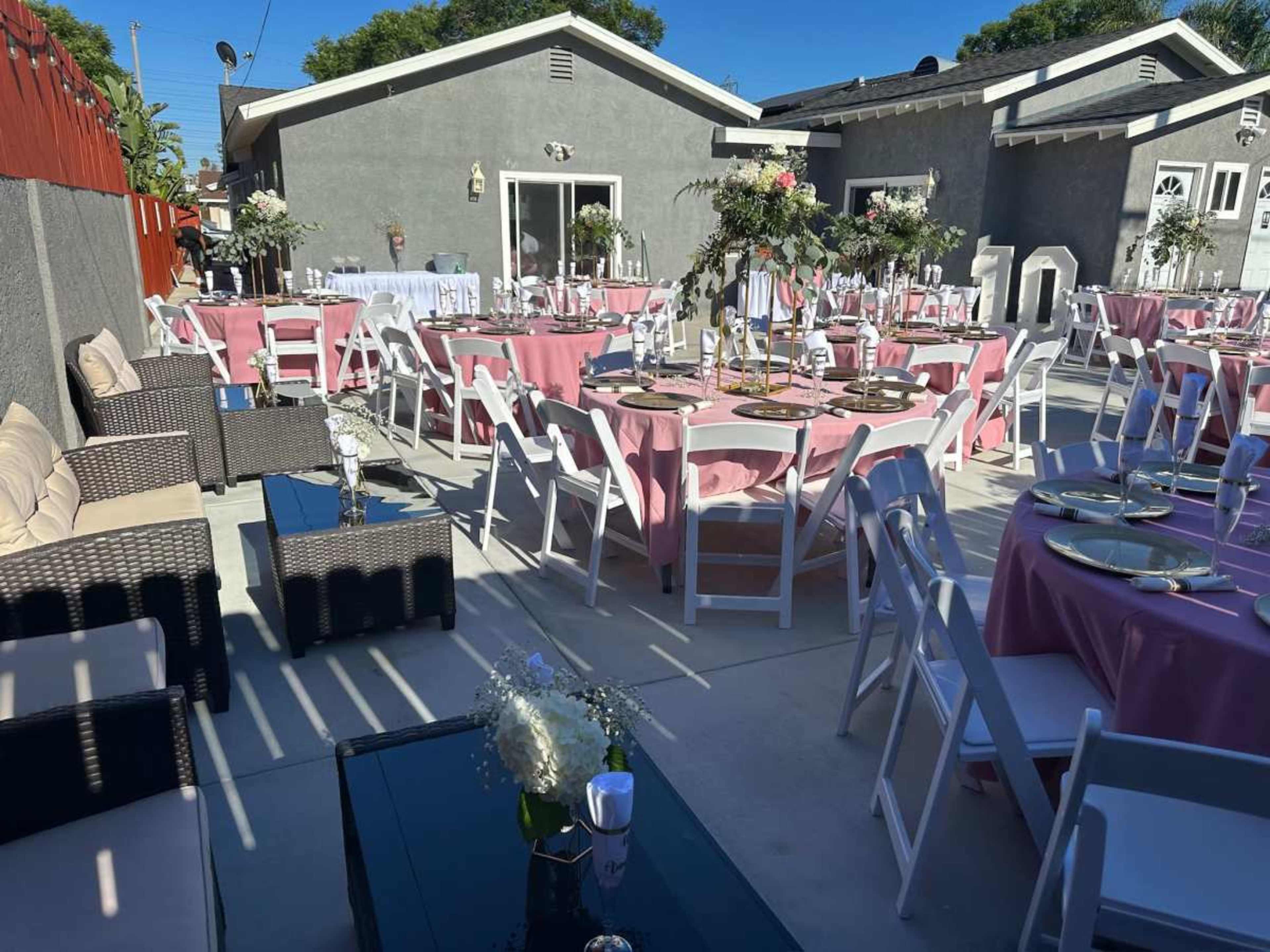 The image shows a decorated outdoor event space with round tables covered in pink tablecloths, white chairs, and floral centerpieces, set against a backdrop of gray buildings.