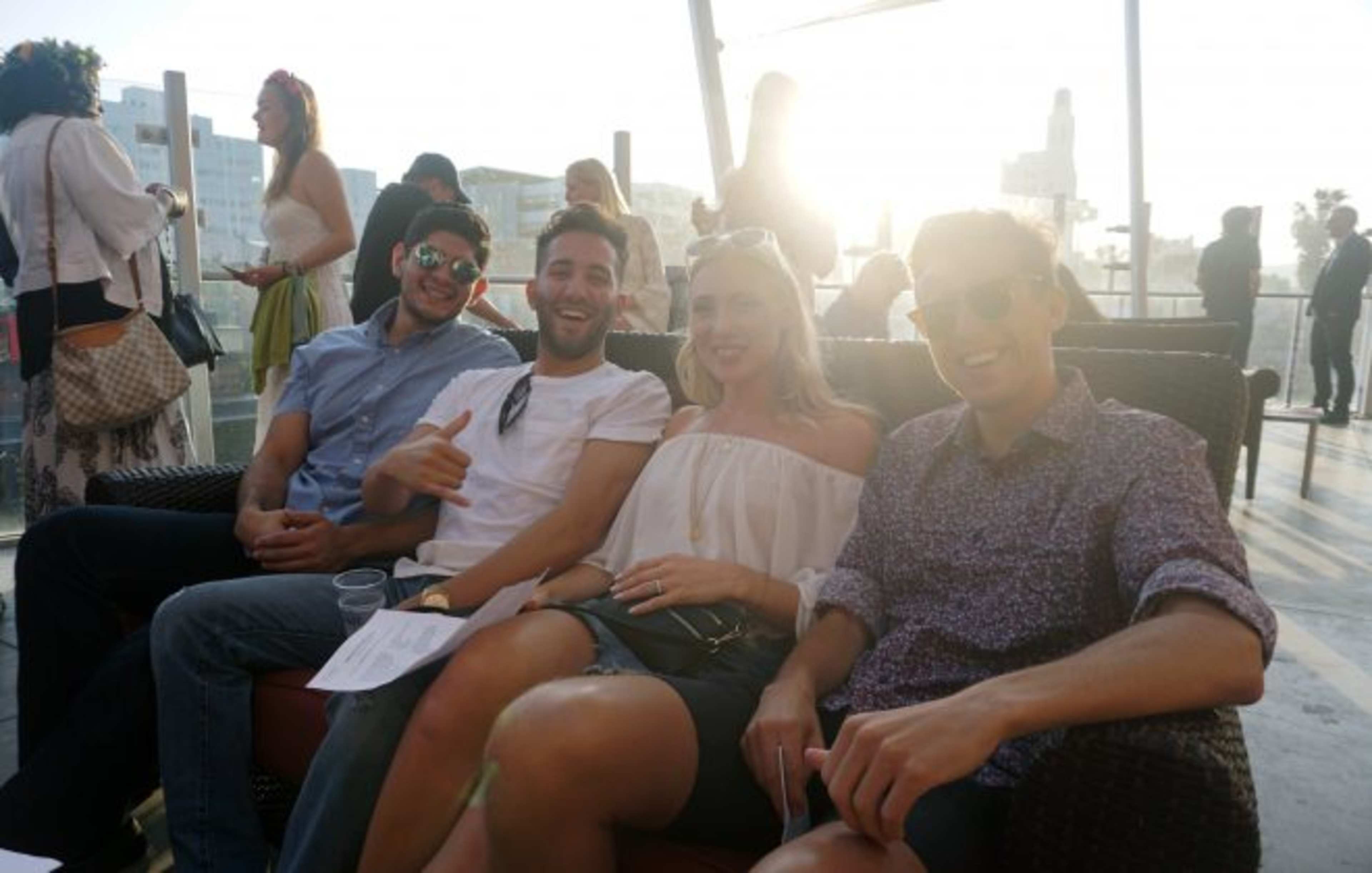 A group of four people sits on a couch, smiling and interacting, with a sunlit background and other people visible in the distance.