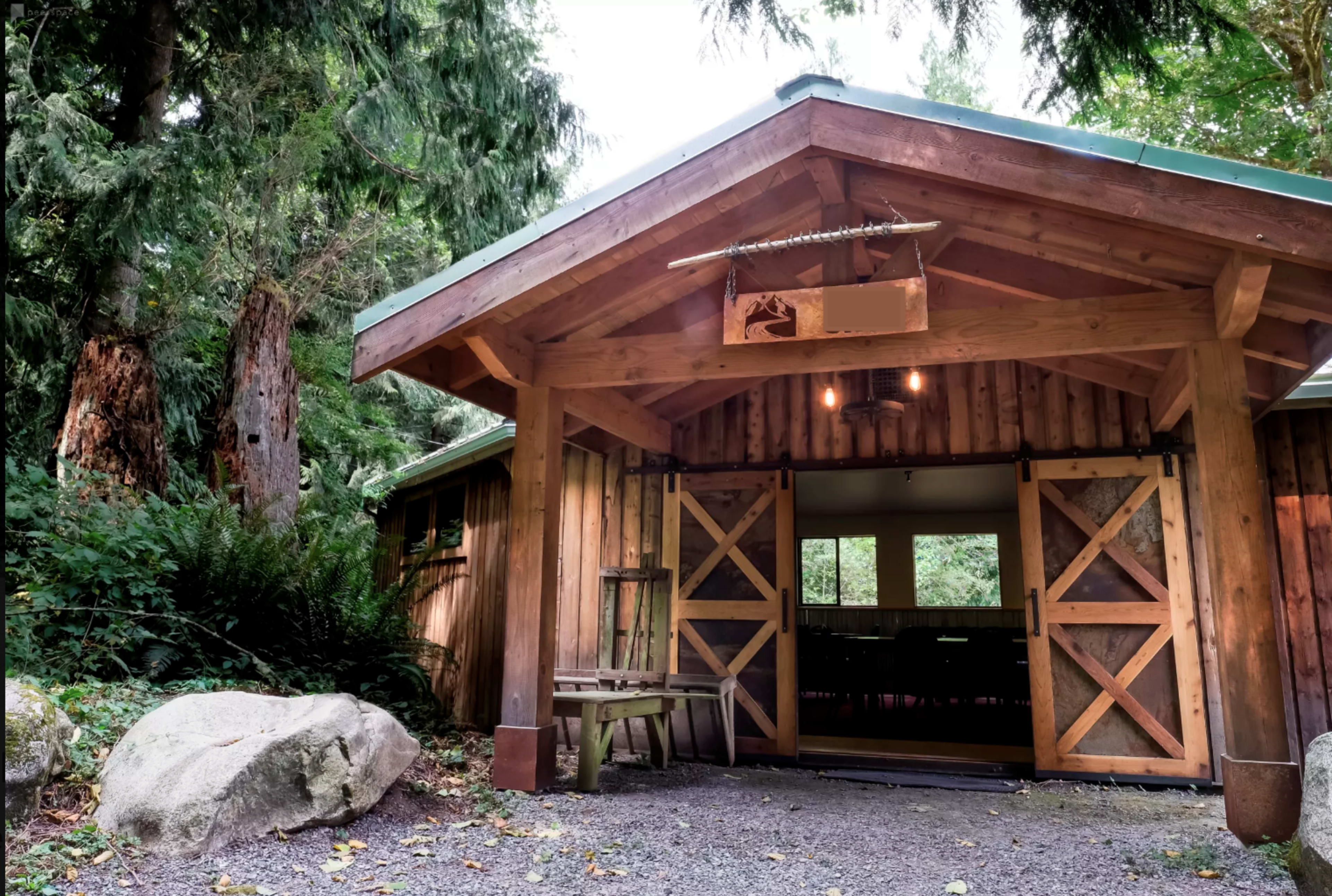A rustic wooden building with a sloped roof and wide double doors, surrounded by trees and gravel pathways.