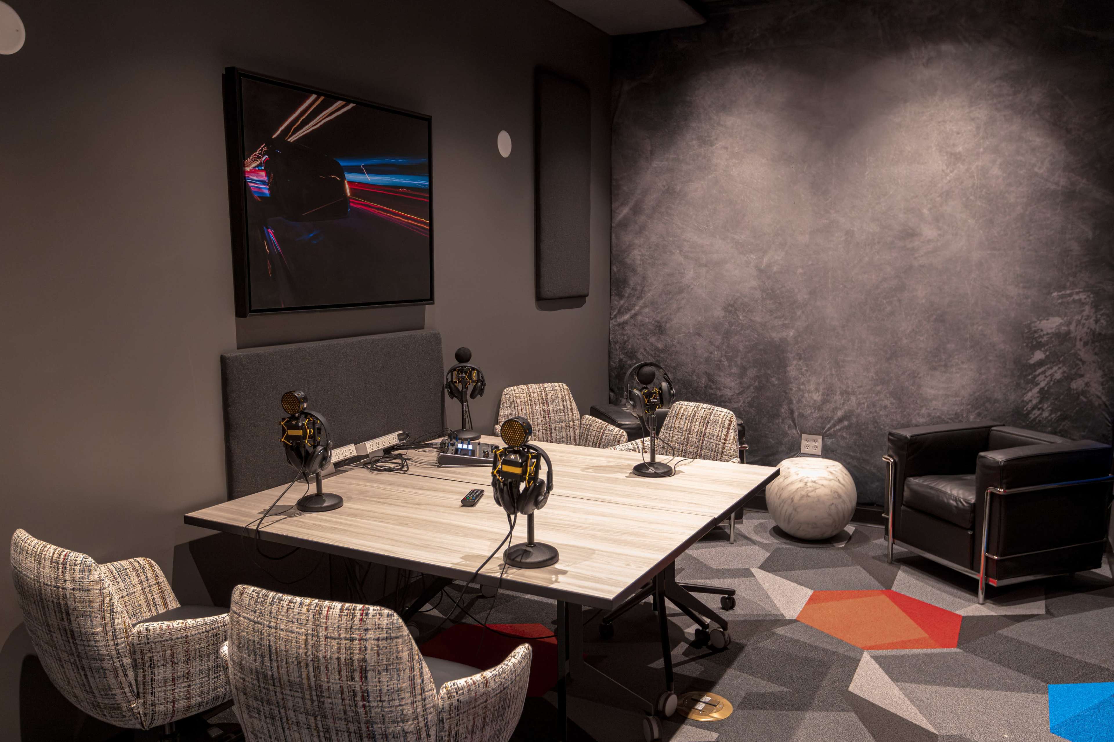 The image shows a modern recording studio with a table, four chairs, microphones, and sound equipment against a textured gray wall.