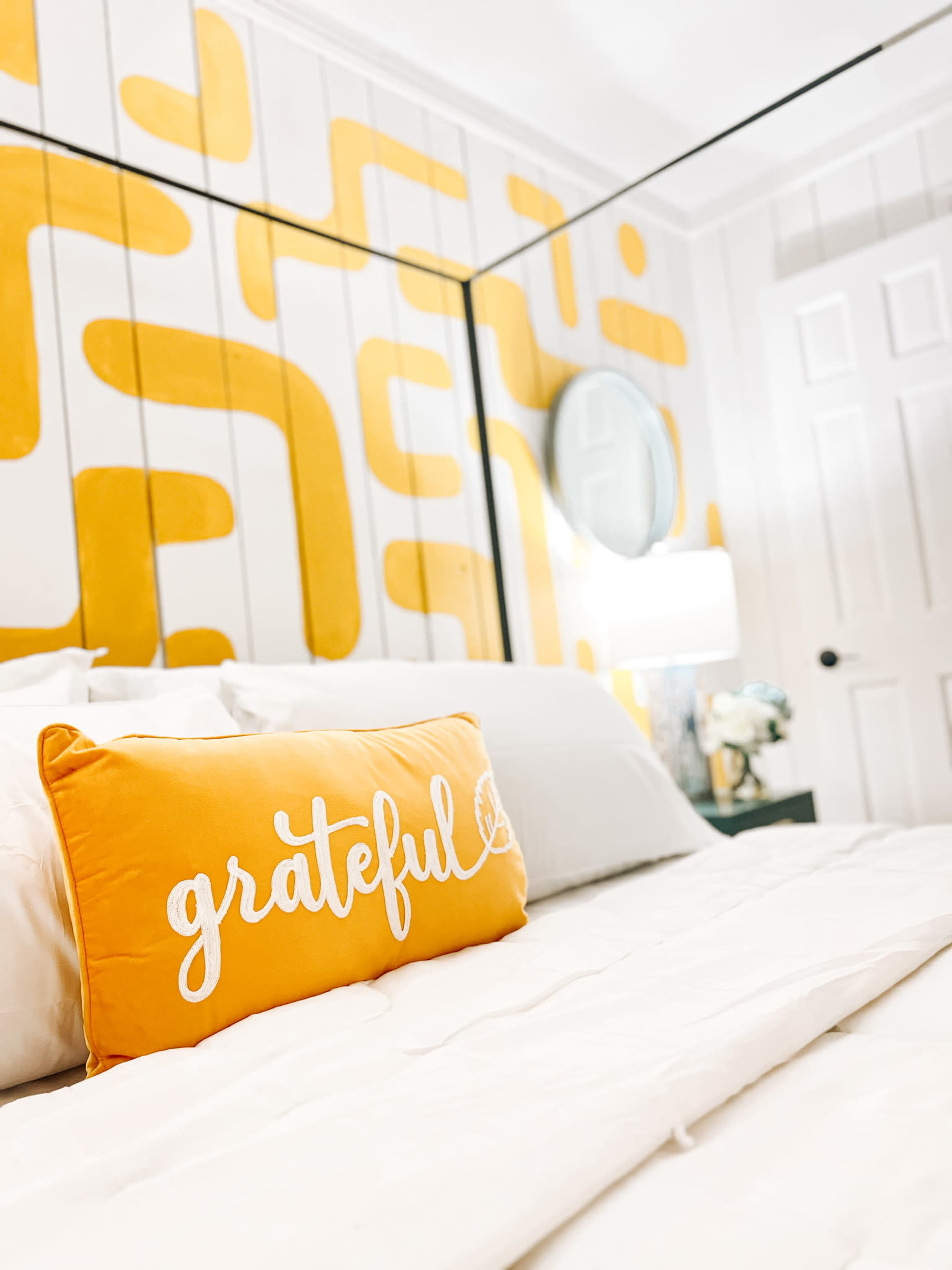 A bed with a white comforter and an orange pillow that has the word "grateful" printed on it, set against a wall featuring a yellow geometric design.