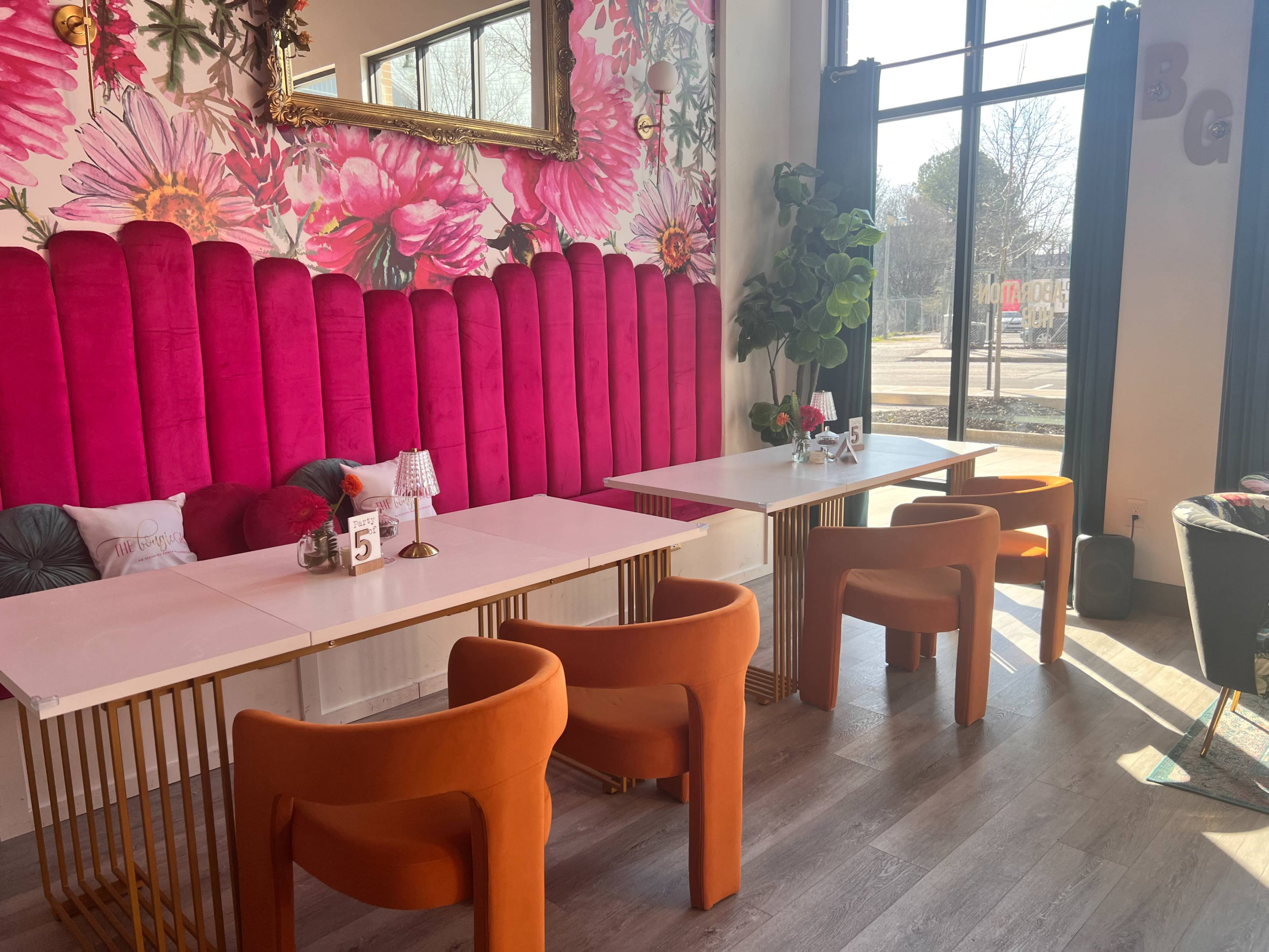 The image shows a bright cafe interior featuring pink upholstered walls, gold metal tables, and orange chairs, with large windows allowing natural light to enter.
