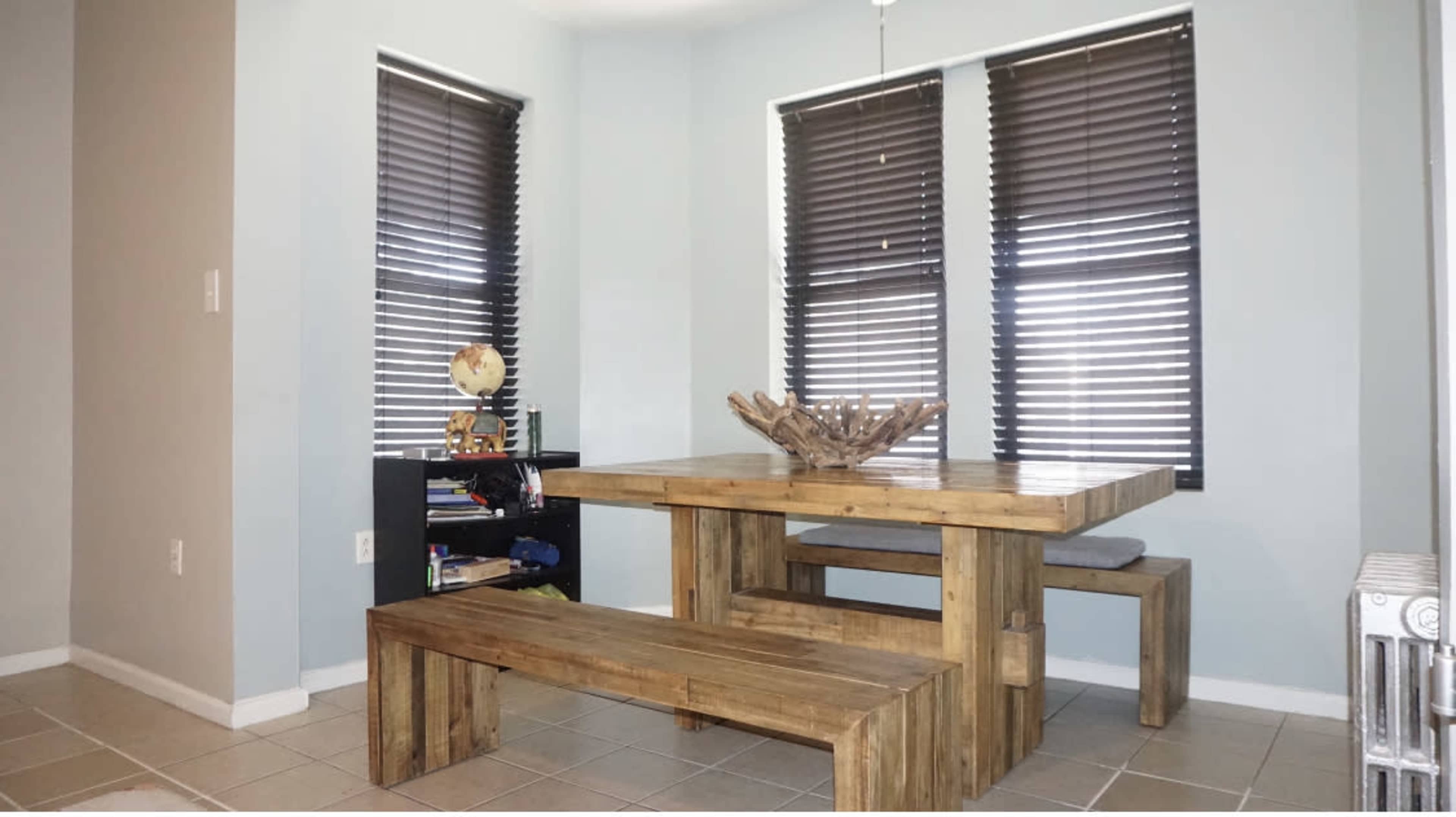 A wooden dining table with matching benches is positioned against walls with two windows covered by slatted blinds in a light blue room.