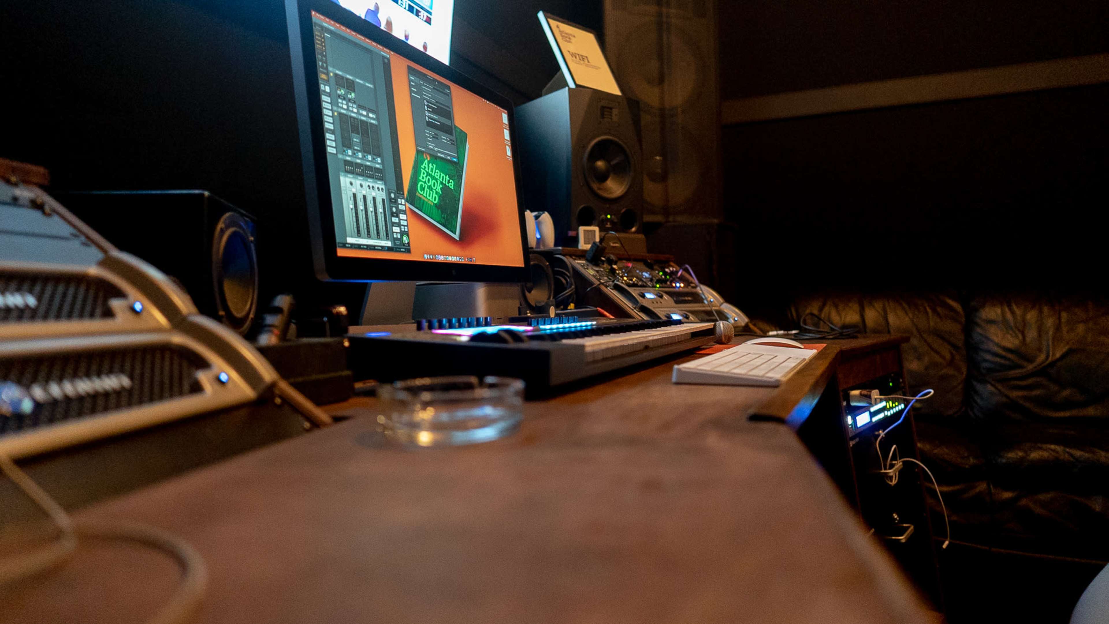 A music production studio features a computer monitor displaying audio software, electronic equipment, and a leather couch in the background.