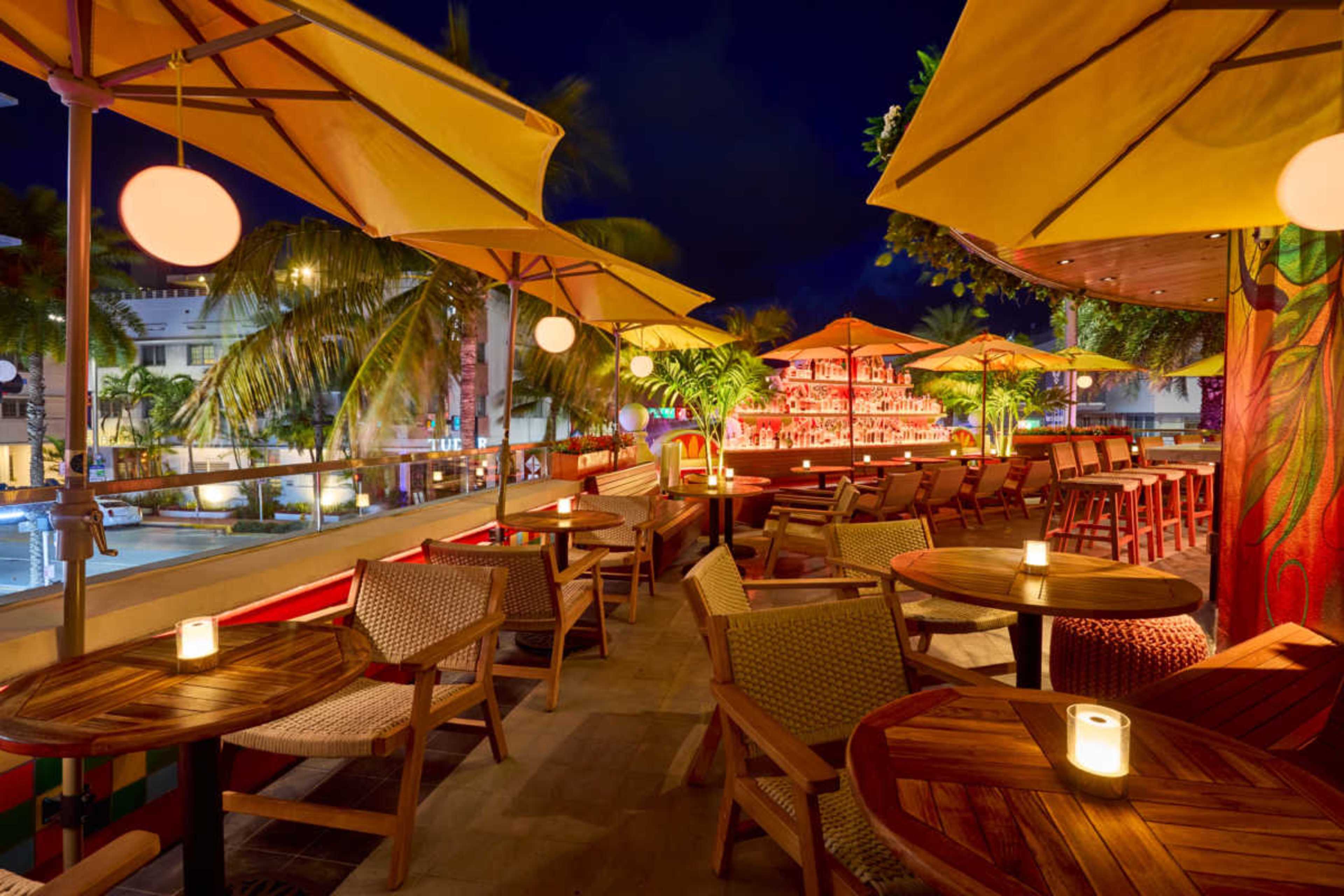 The image shows a vibrant outdoor seating area of a restaurant or bar with wooden tables and chairs, yellow umbrellas, and a brightly lit bar in the background.
