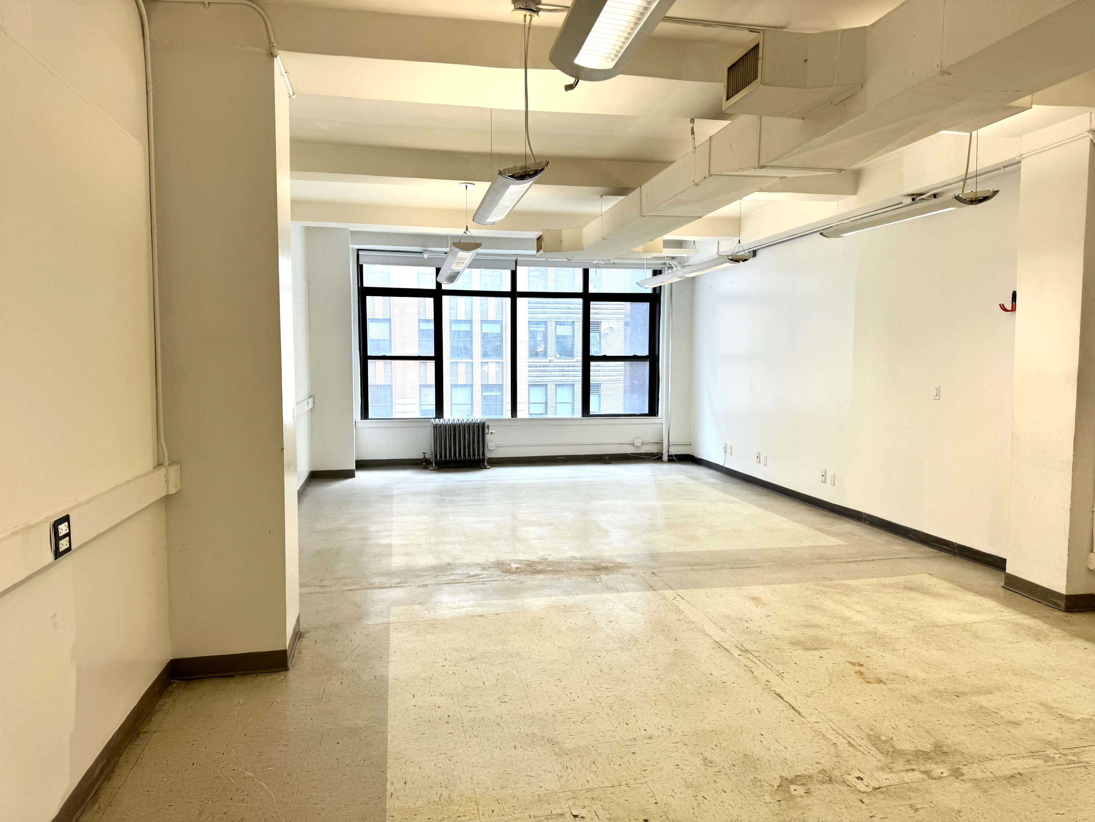 The image shows a vacant, sparsely furnished room with large windows and exposed ceiling beams.