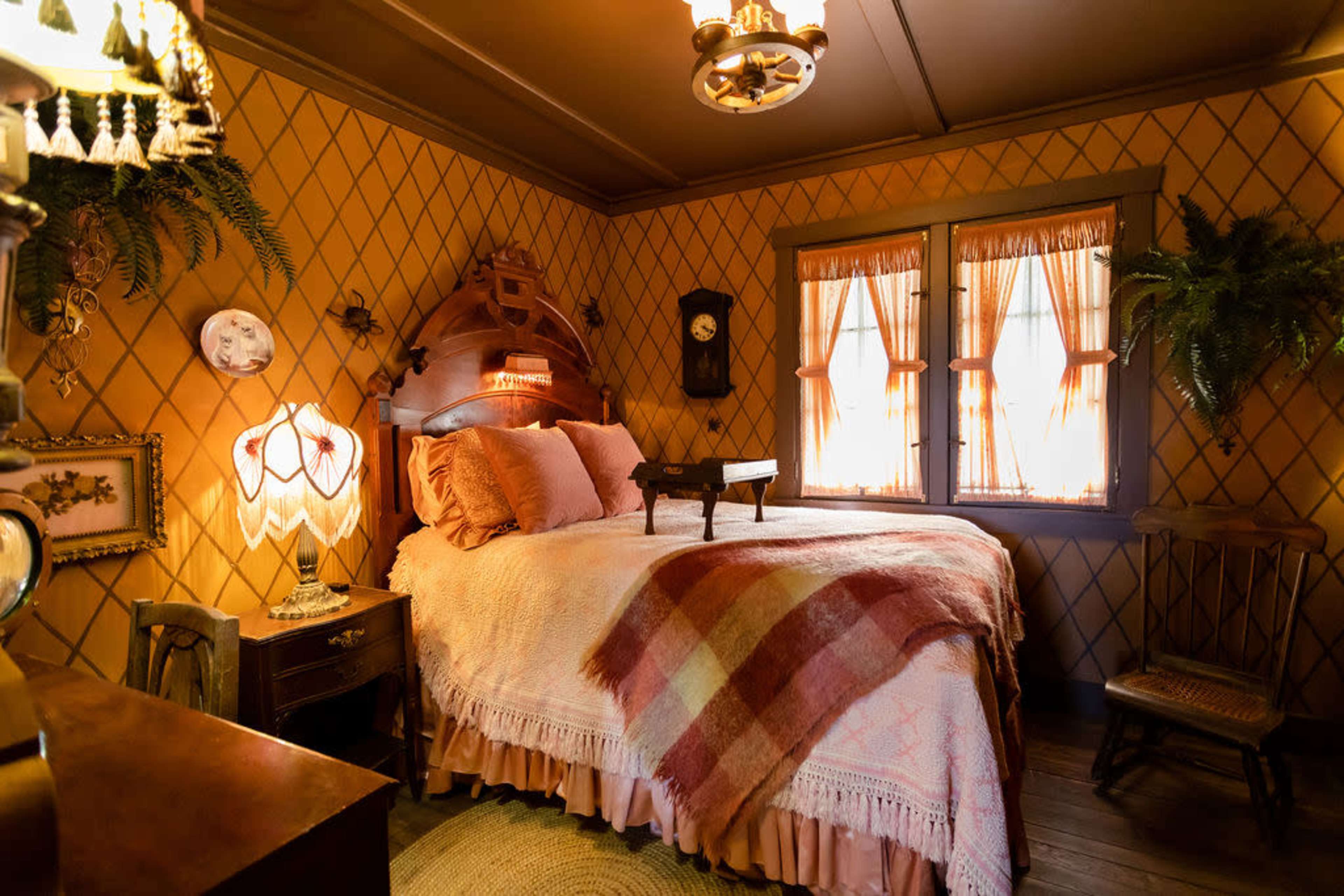 The room features a bed with a patterned blanket, two windows with sheer curtains, and vintage decor elements such as a chandelier and wall-mounted plants.