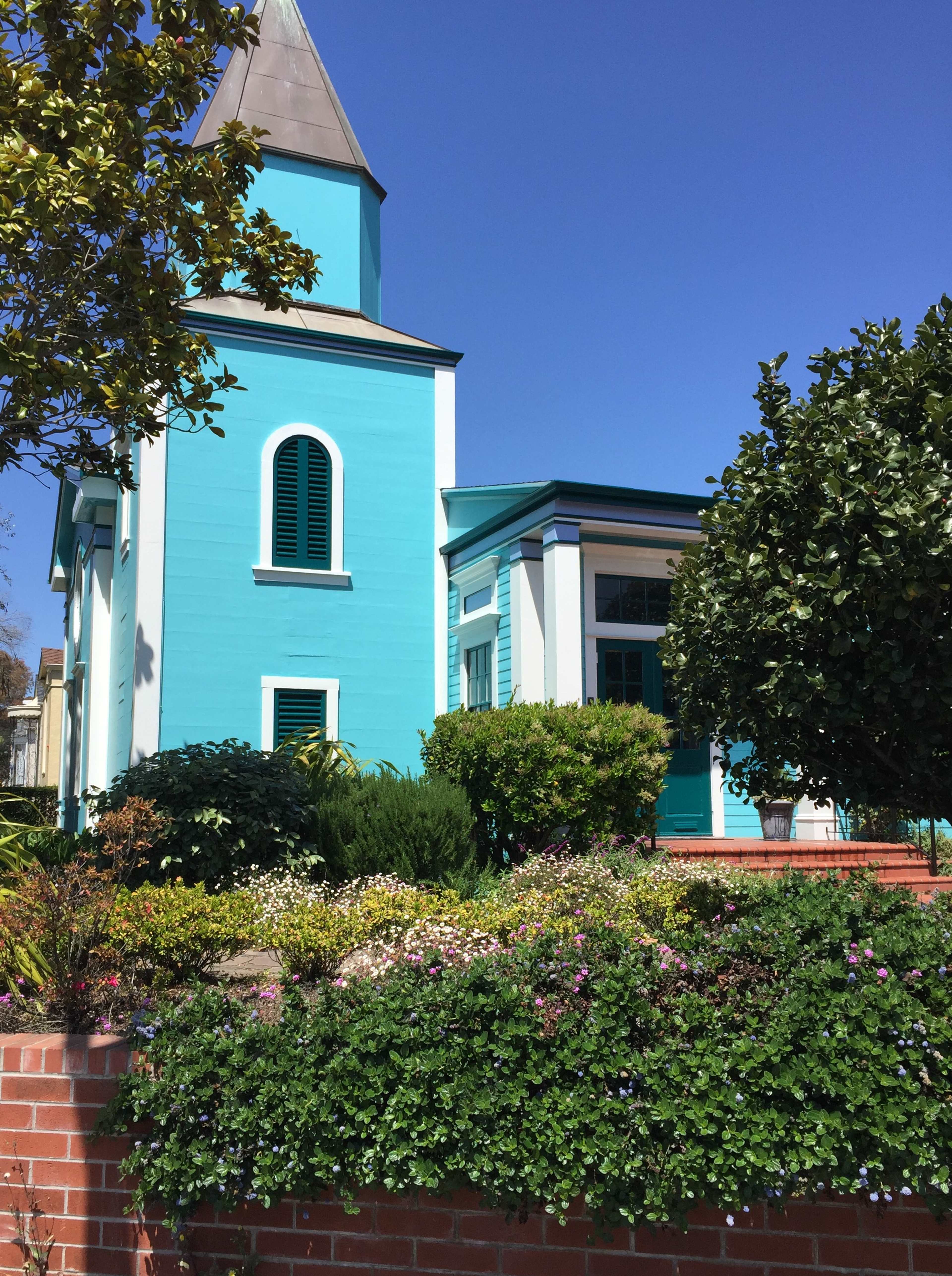 A turquoise building with a pointed roof, surrounded by green plants and colorful flowers.