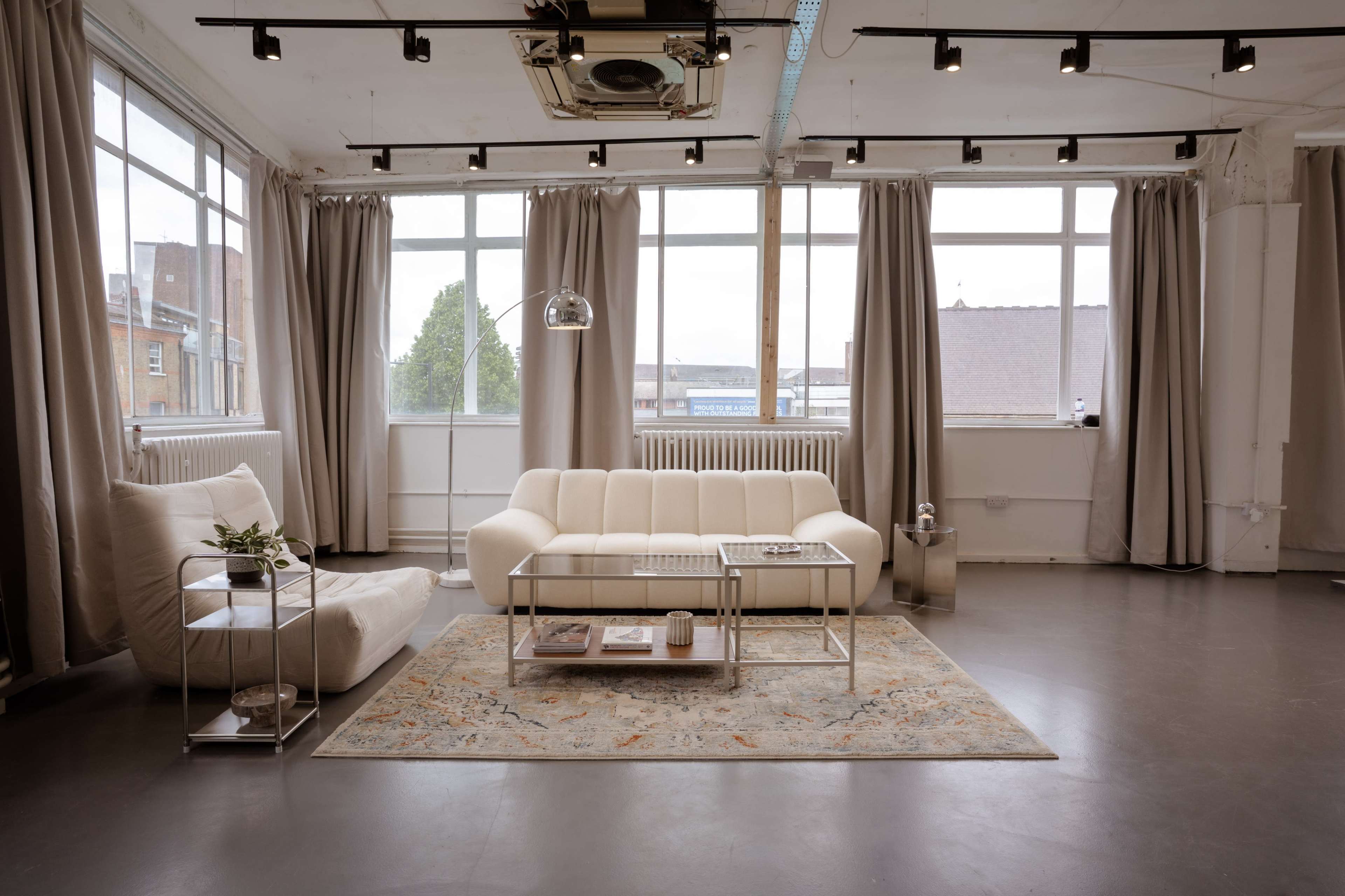 The image shows a modern, minimalist living room with large windows, a white sofa, a decorative area rug, and light curtains.