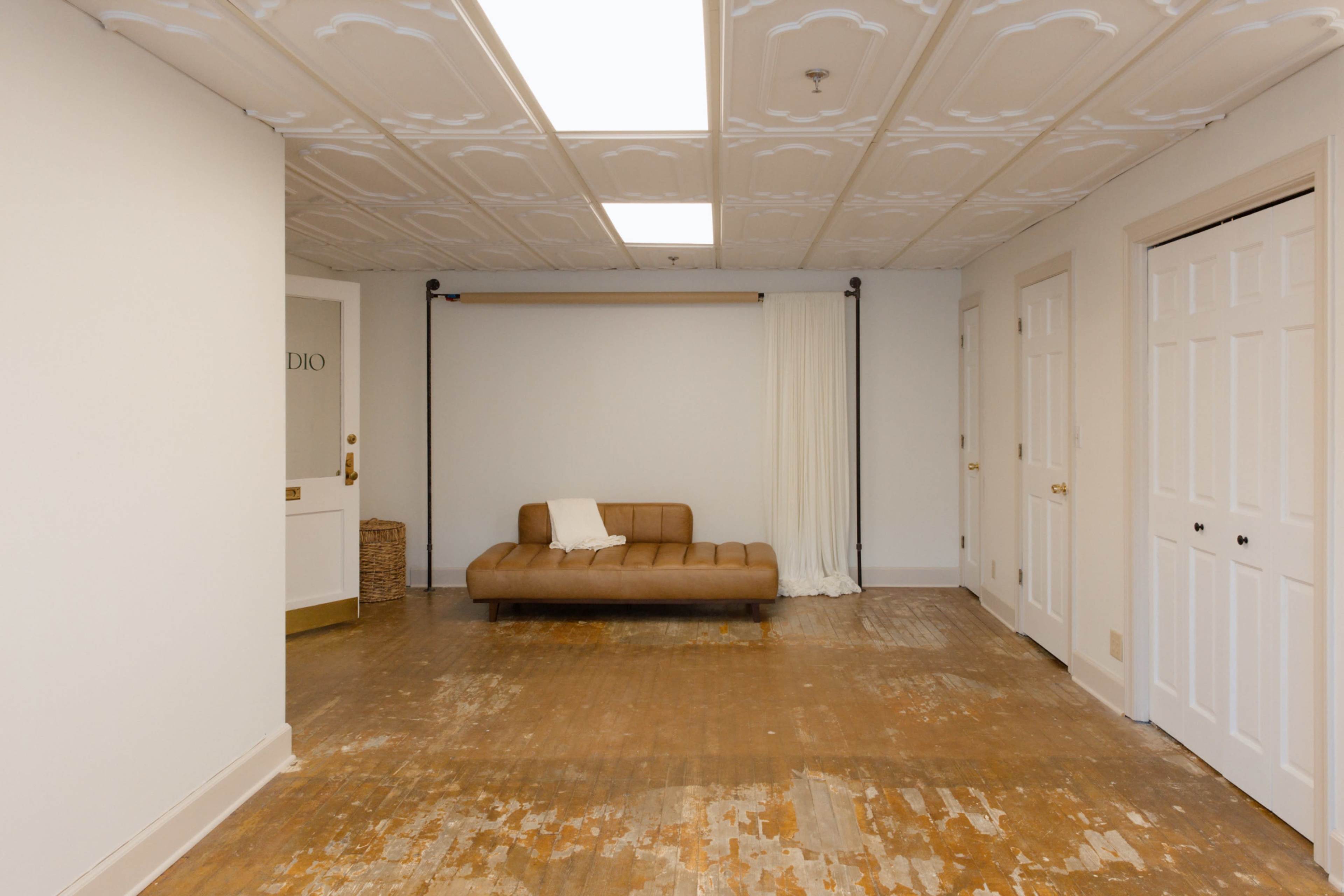 The image shows a spacious, empty room with a brown couch against one wall, white curtains, and double doors.