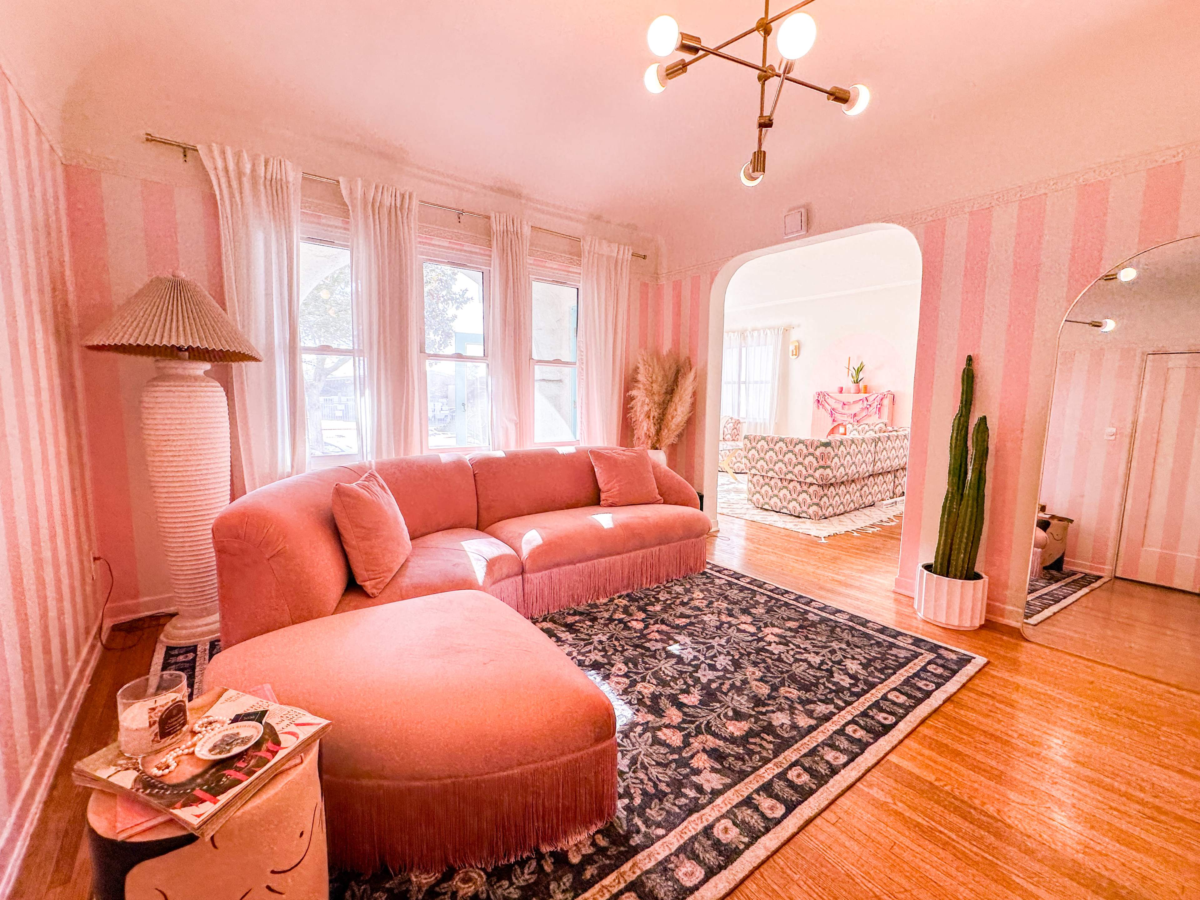 The living room features a pink sectional sofa, a patterned rug, and large windows with sheer curtains, complemented by a decorative lamp and a potted cactus.