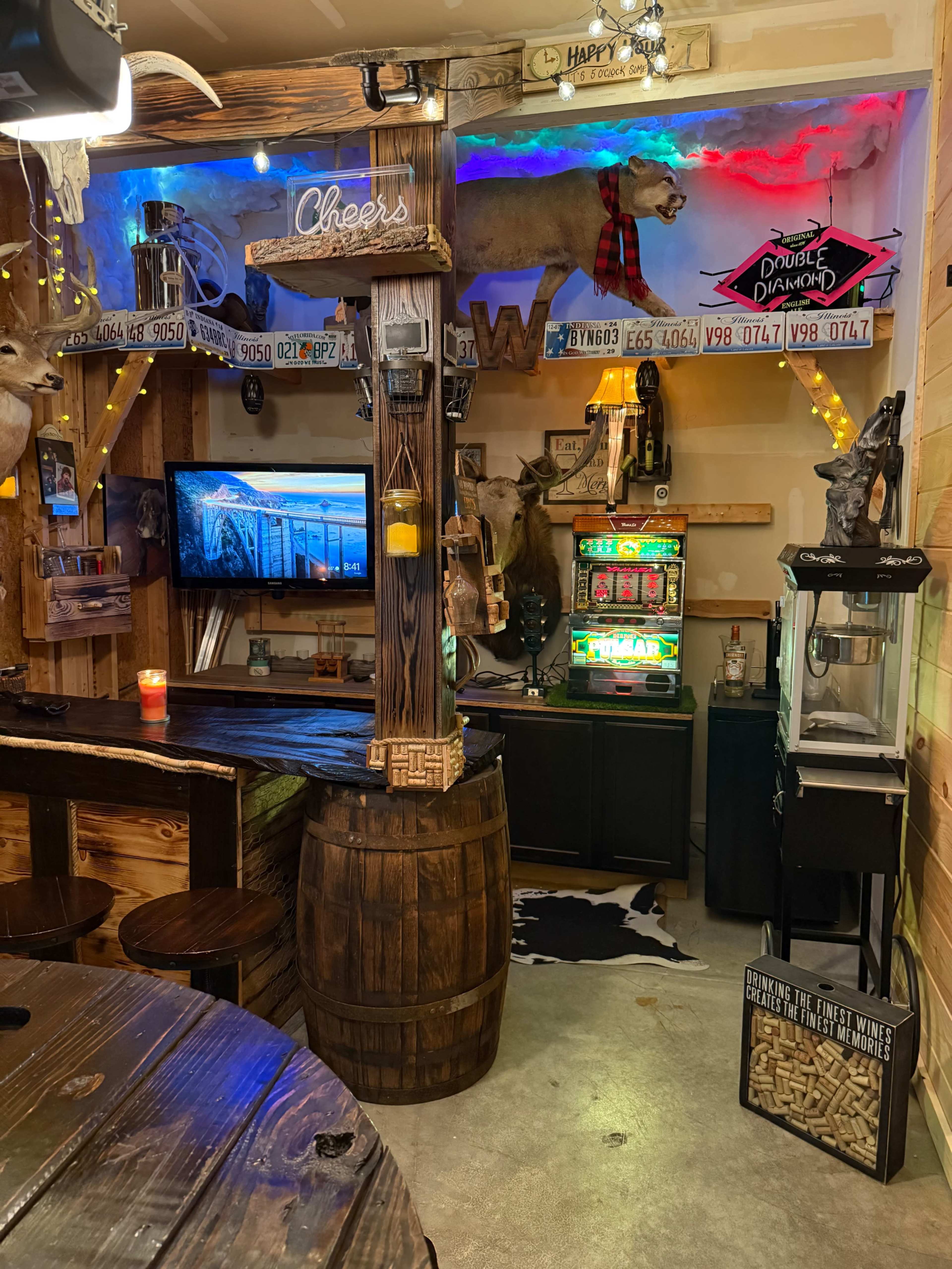 The image shows a rustic home bar setup featuring a wooden counter, a gaming machine, mounted hunting trophies, and ambient lighting.