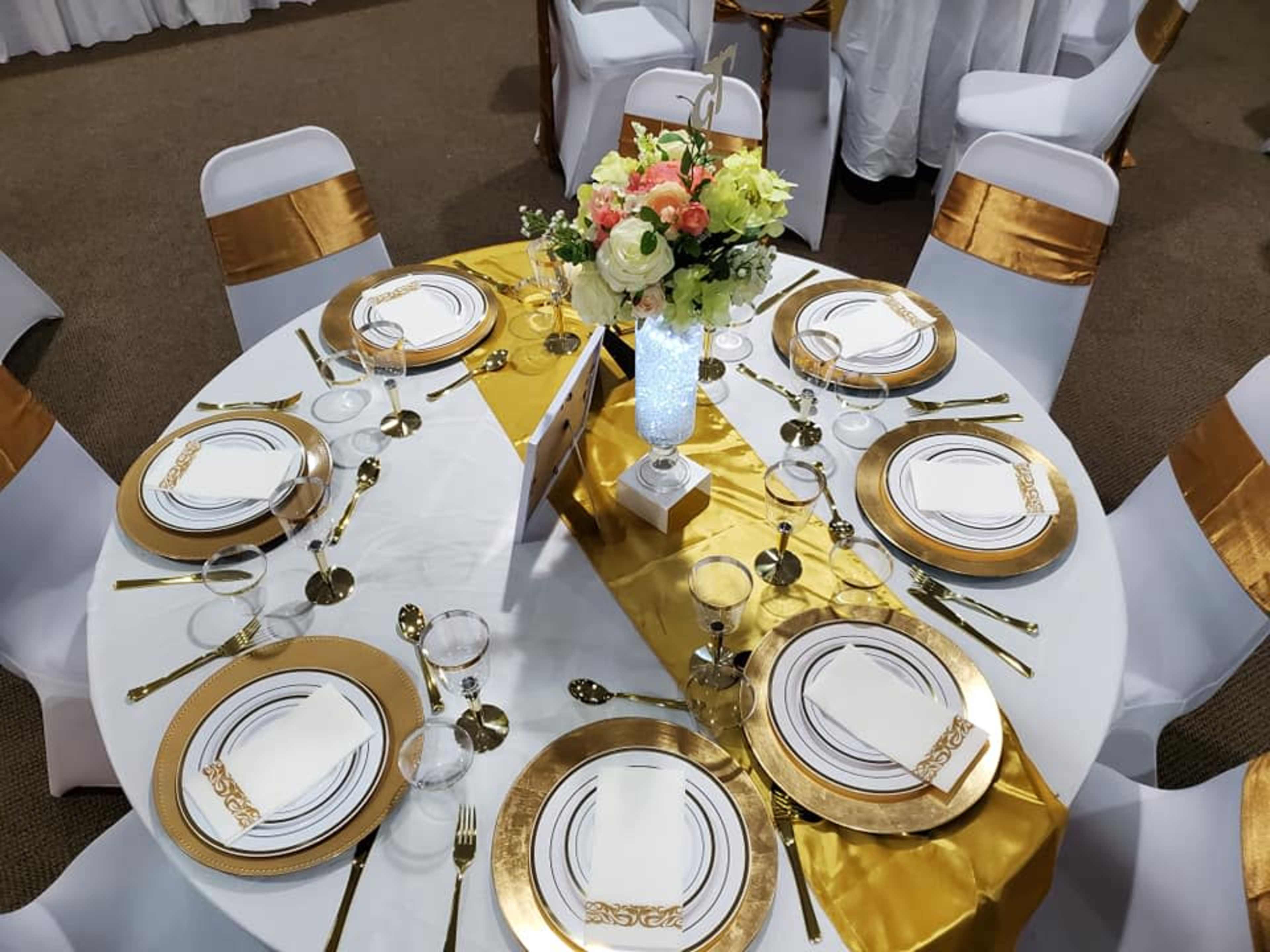 The image shows a round dining table set with white tablecloths, gold-accented plates, glasses, and floral arrangements in a decorative vase.
