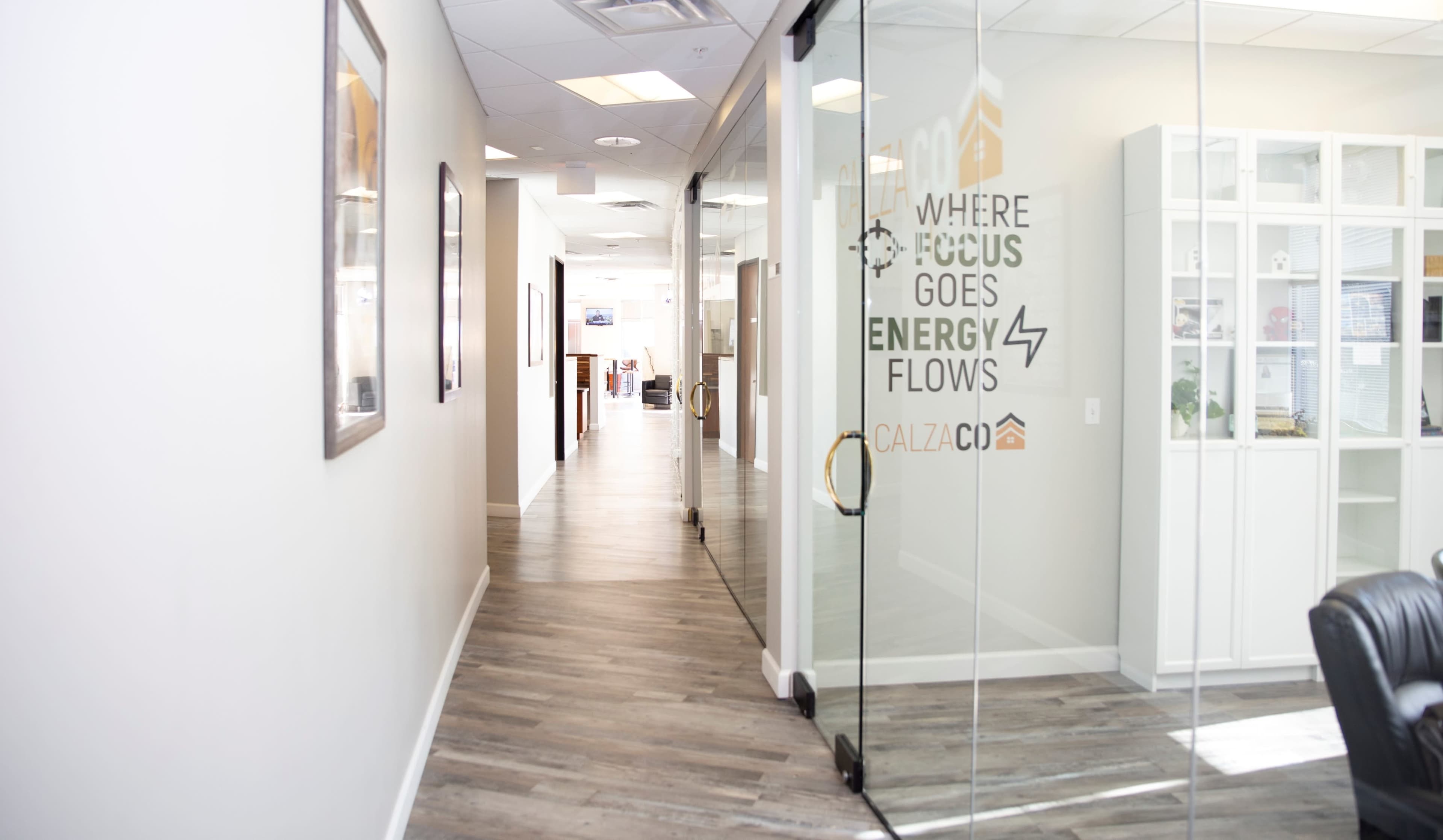 A long office corridor features glass doors with a motivational slogan and wooden flooring leading to various workspaces.