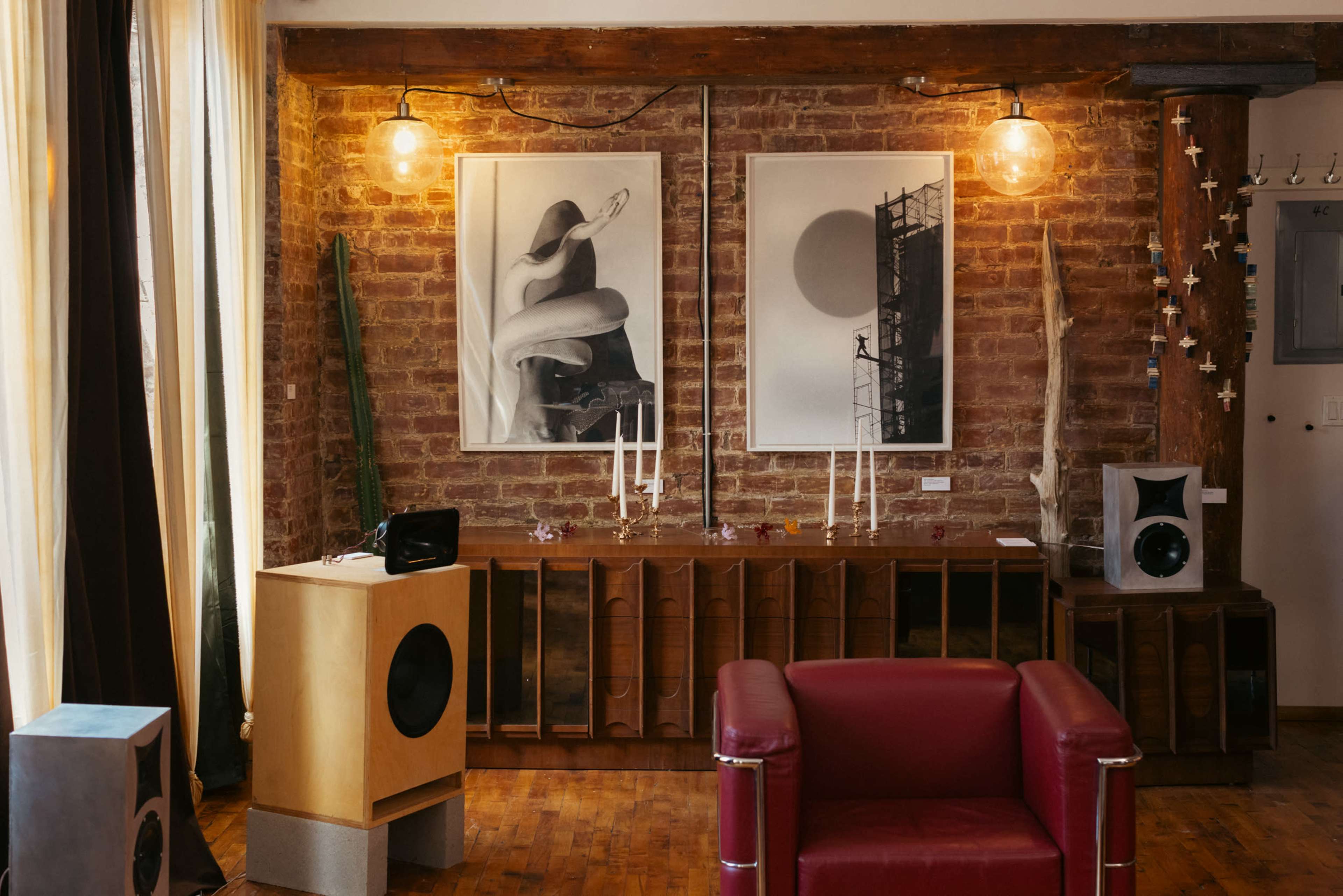 The image shows a modern interior featuring a brick wall, abstract artwork on display, a wooden cabinet, a red armchair, and various decorative elements.