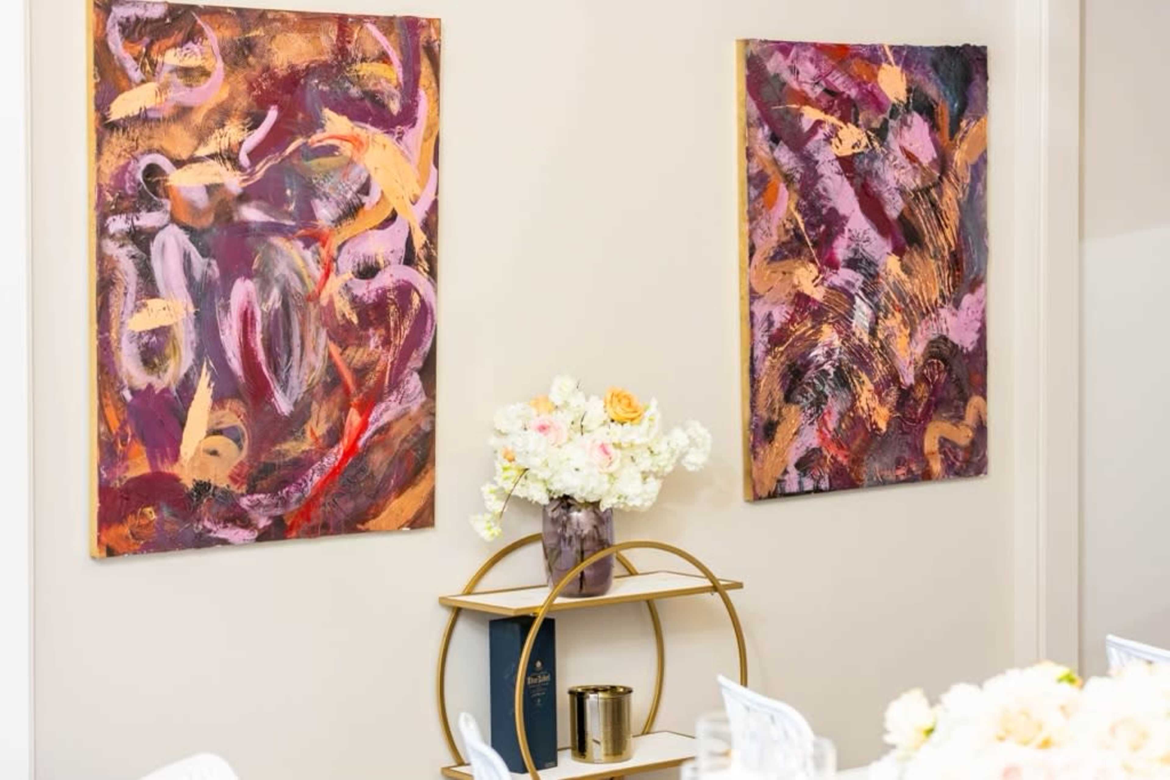 Two abstract paintings hang on a wall next to a small round shelf with a vase of flowers and a book.