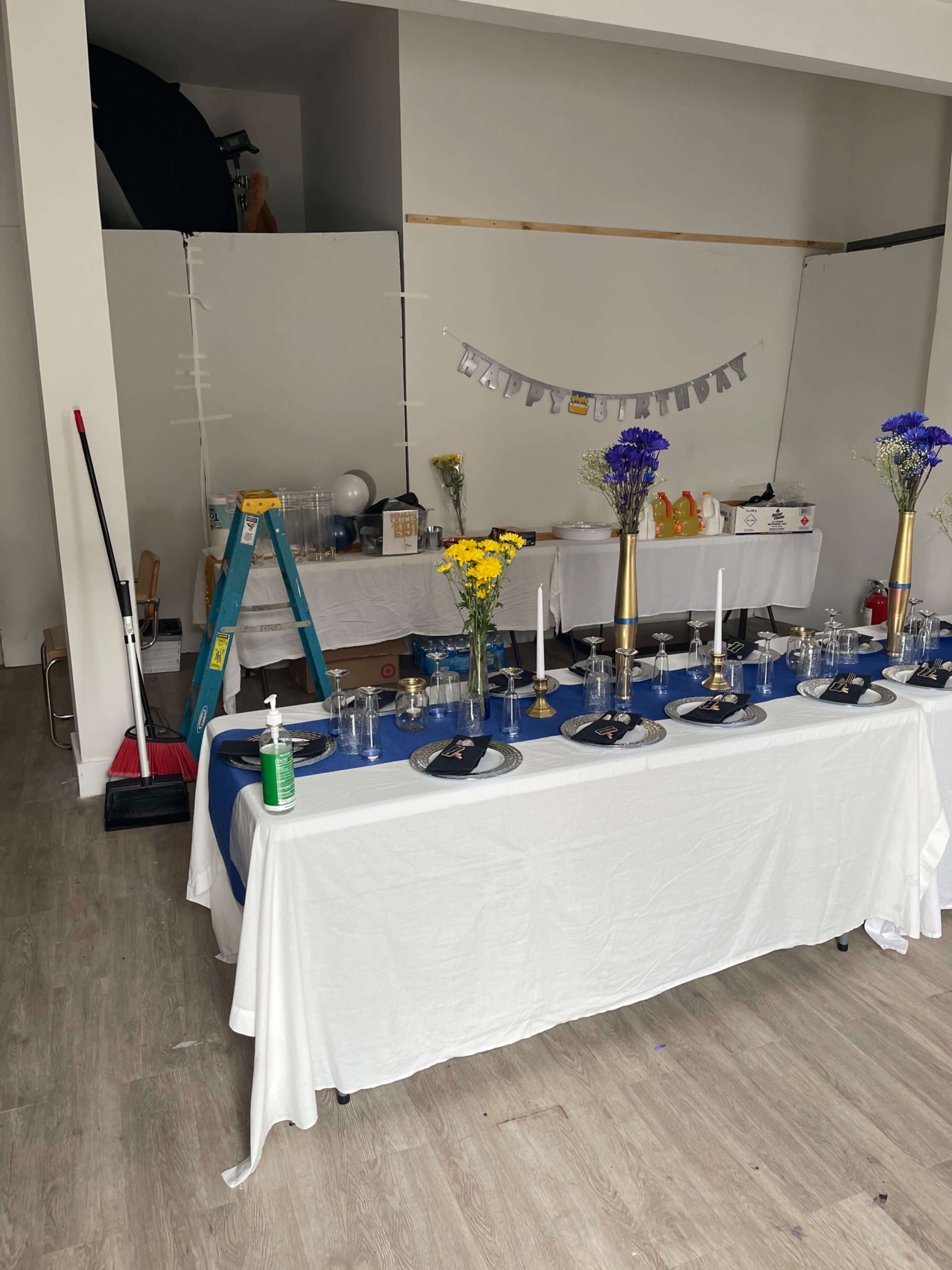 A decorated table with white tablecloths and blue accents is set up for a birthday celebration in a room with a ladder and cleaning supplies visible in the background.