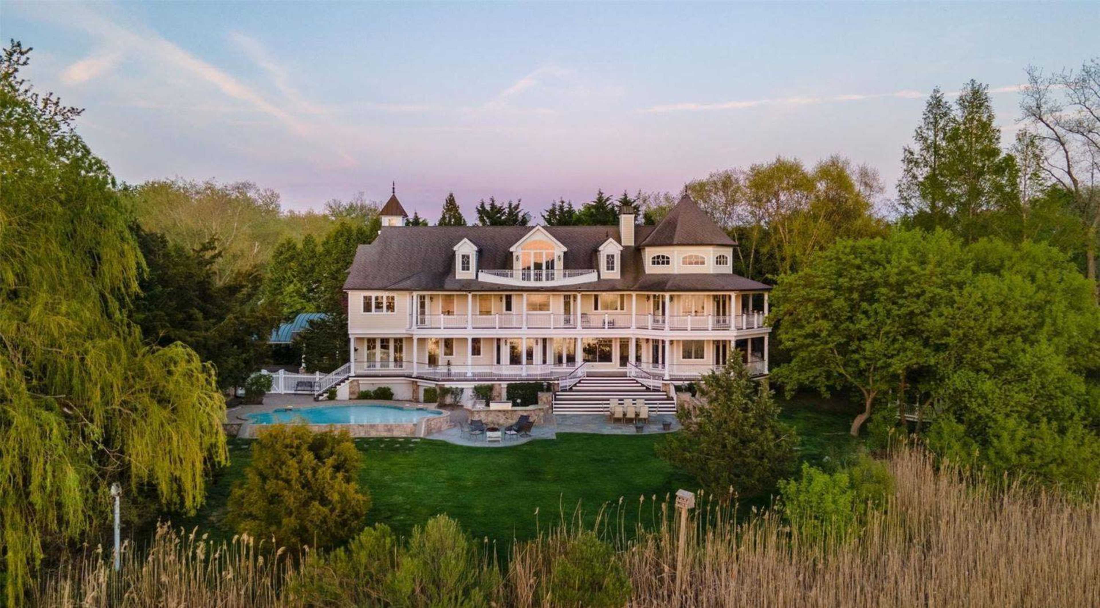 A large, multi-level waterfront house features several balconies, a pool in the backyard, and is surrounded by lush greenery.