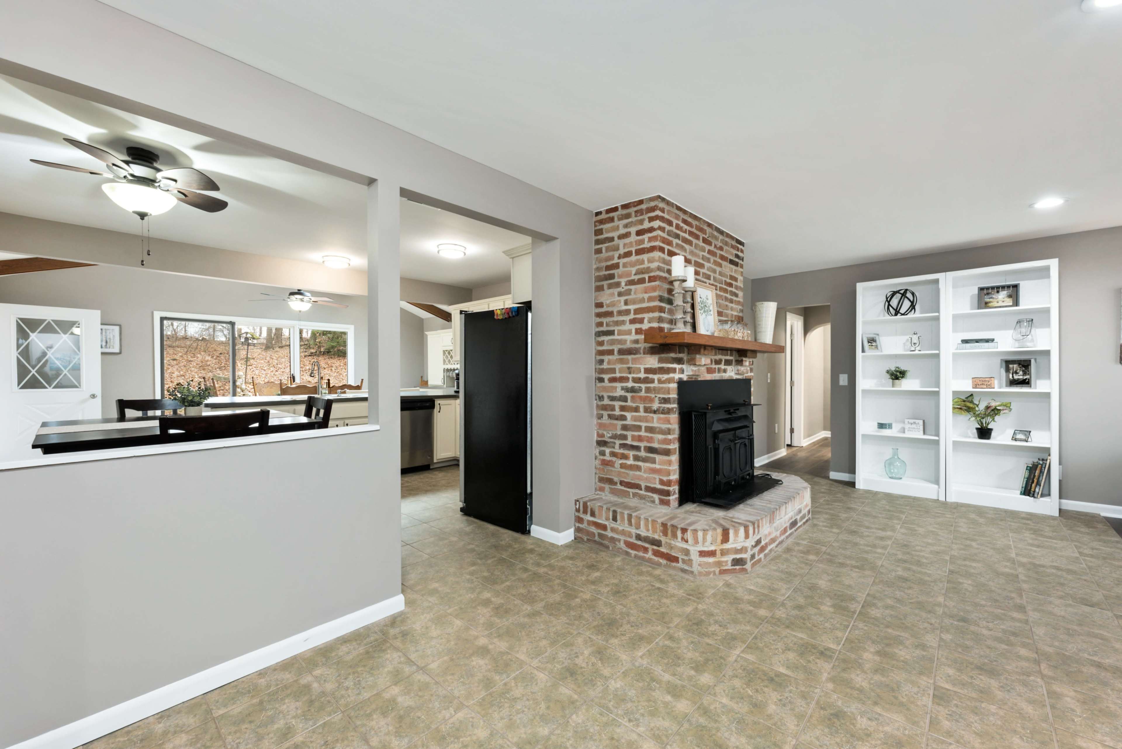 The image shows an interior living space featuring a brick fireplace with a wooden mantel, built-in shelves, and a view into a kitchen area through an open wall.