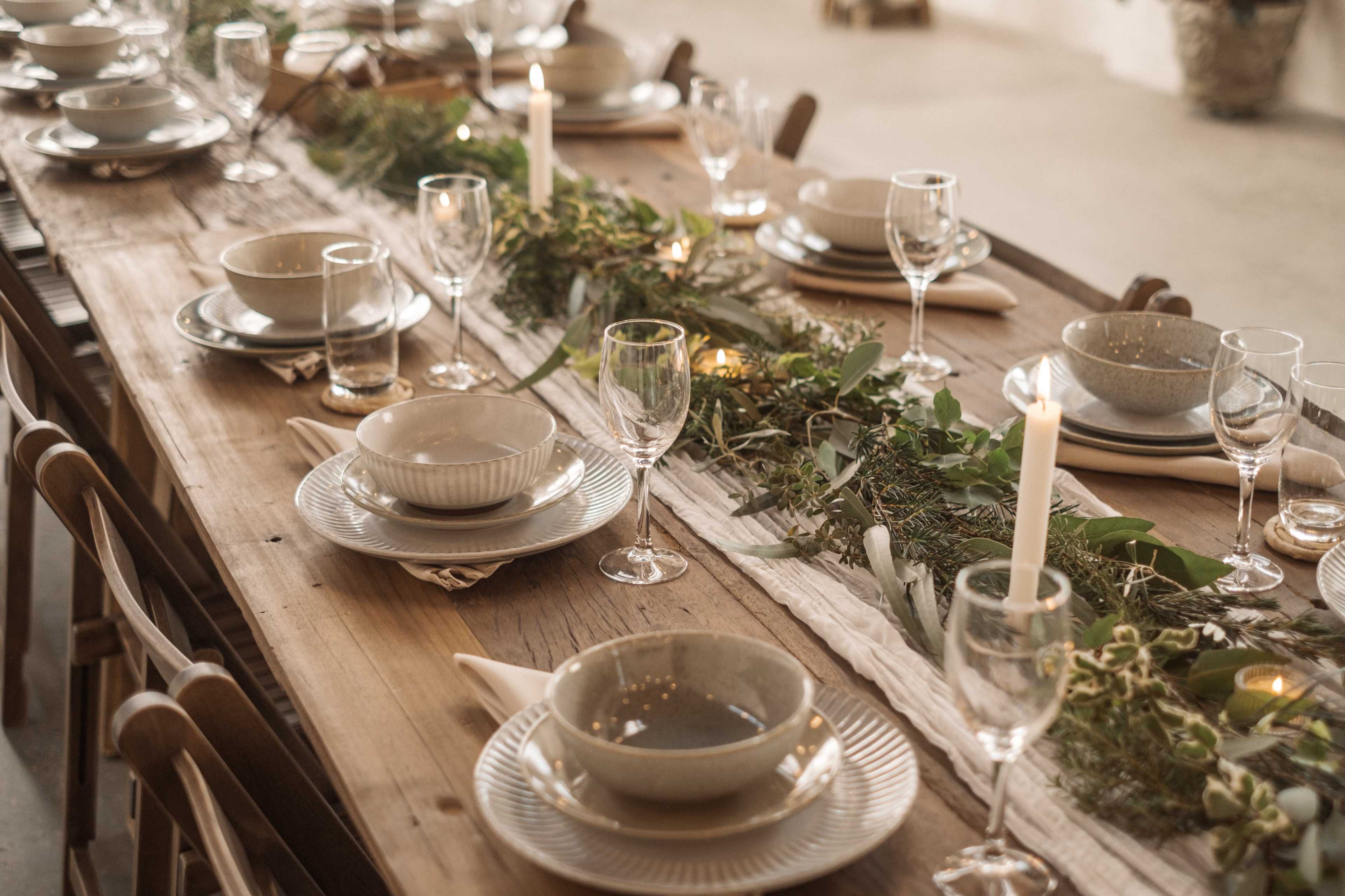 A long wooden table is set for a meal with plates, bowls, glasses, and candles, surrounded by greenery.