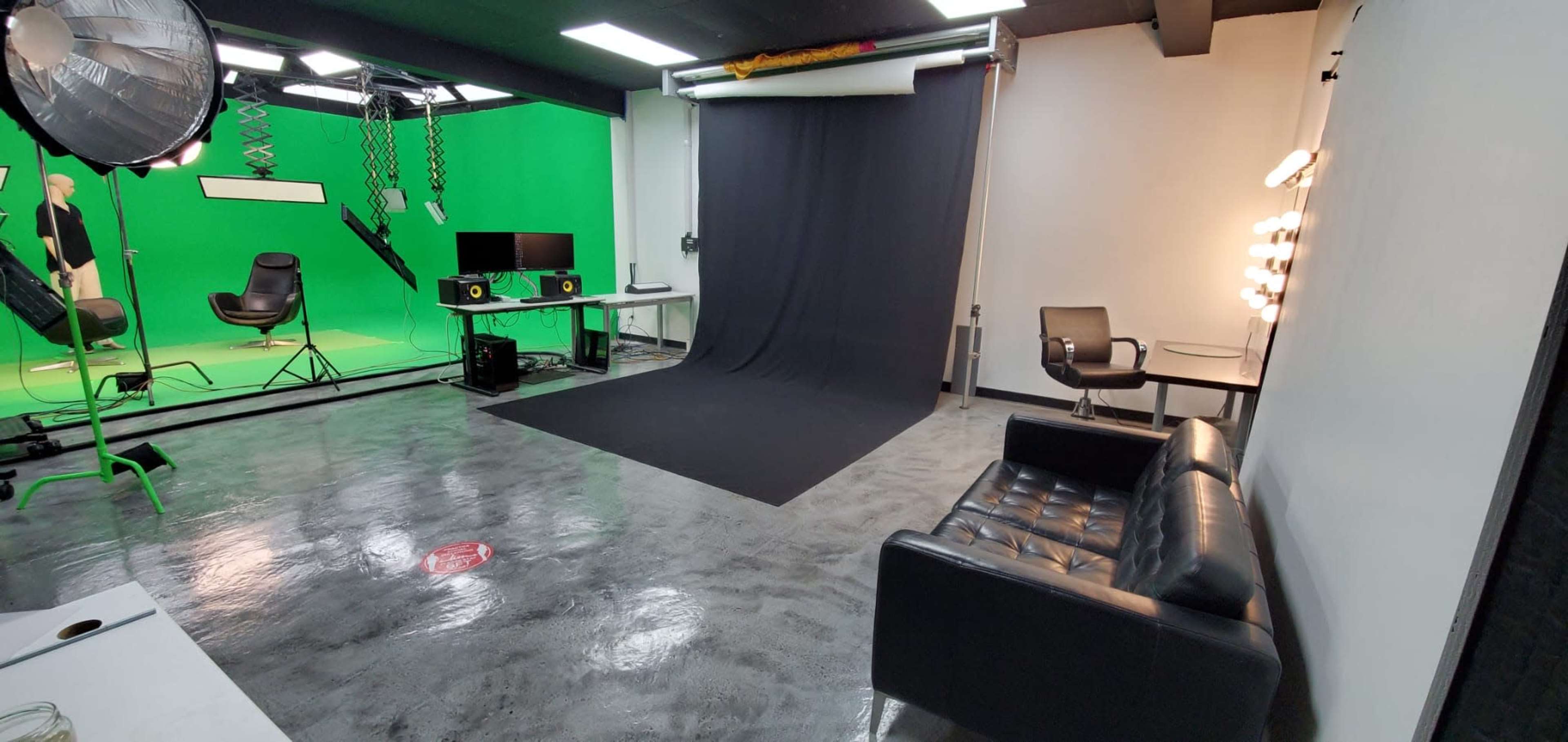 The image shows a video production studio featuring green screens, a black backdrop, and a seating area with a black leather couch.