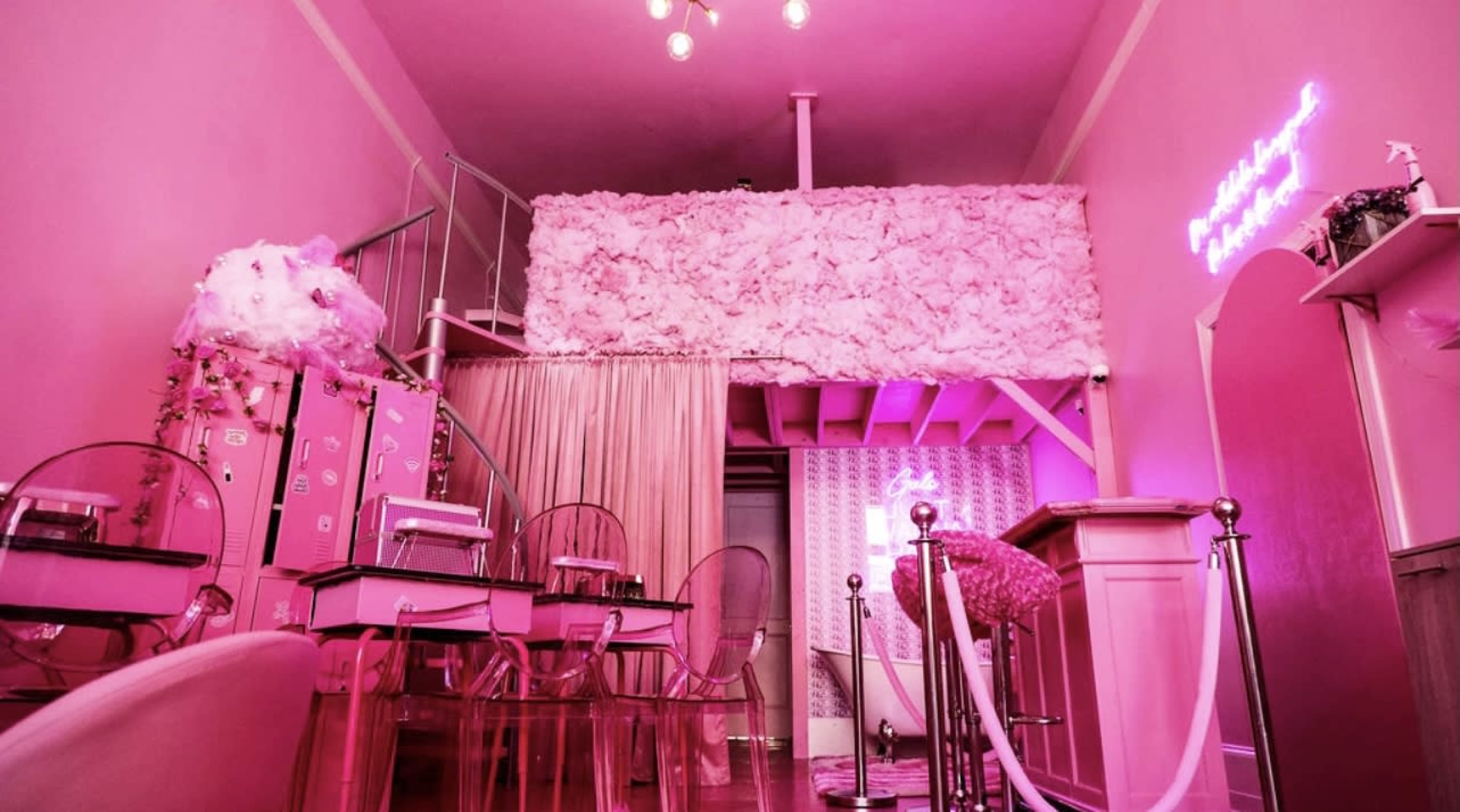 A vibrant, pink-themed café interior with floral decor, transparent chairs, and neon signage.