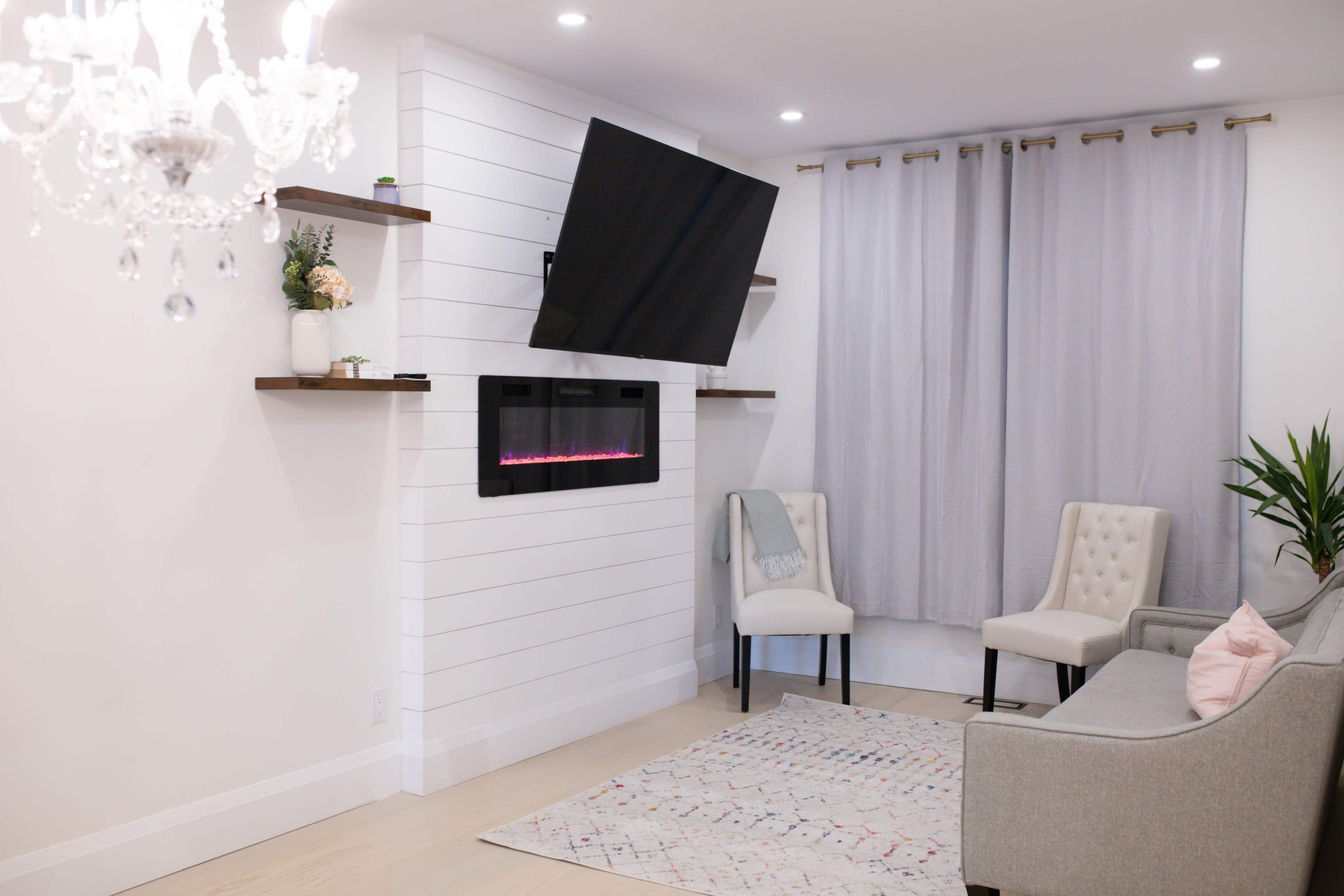 A modern living room features a wall-mounted TV above a sleek fireplace, flanked by shelves, with two chairs, a sofa, and light gray curtains.